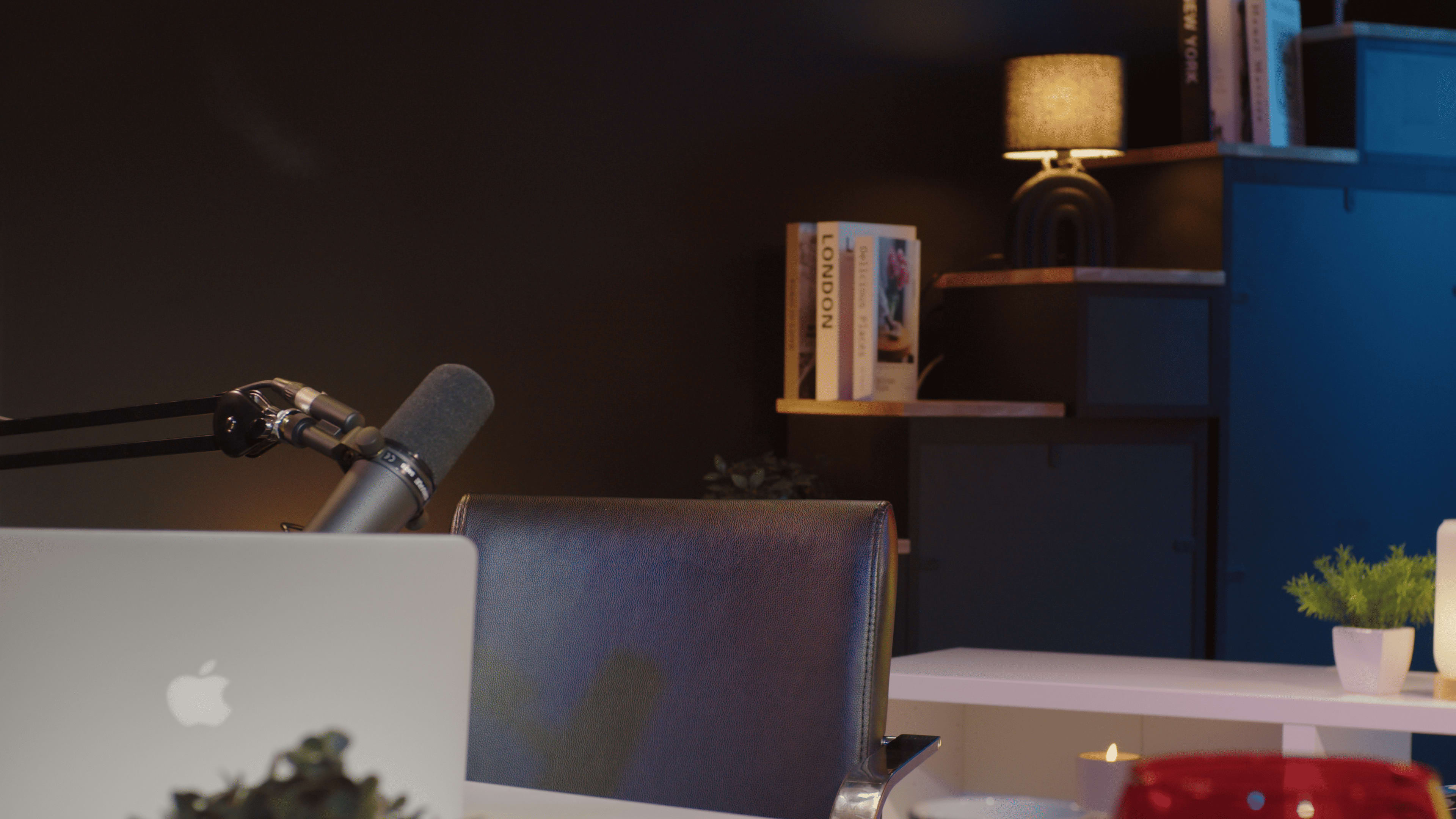 A modern office space features a microphone on a desk next to a laptop, with shelves holding books and a lamp illuminating the area.