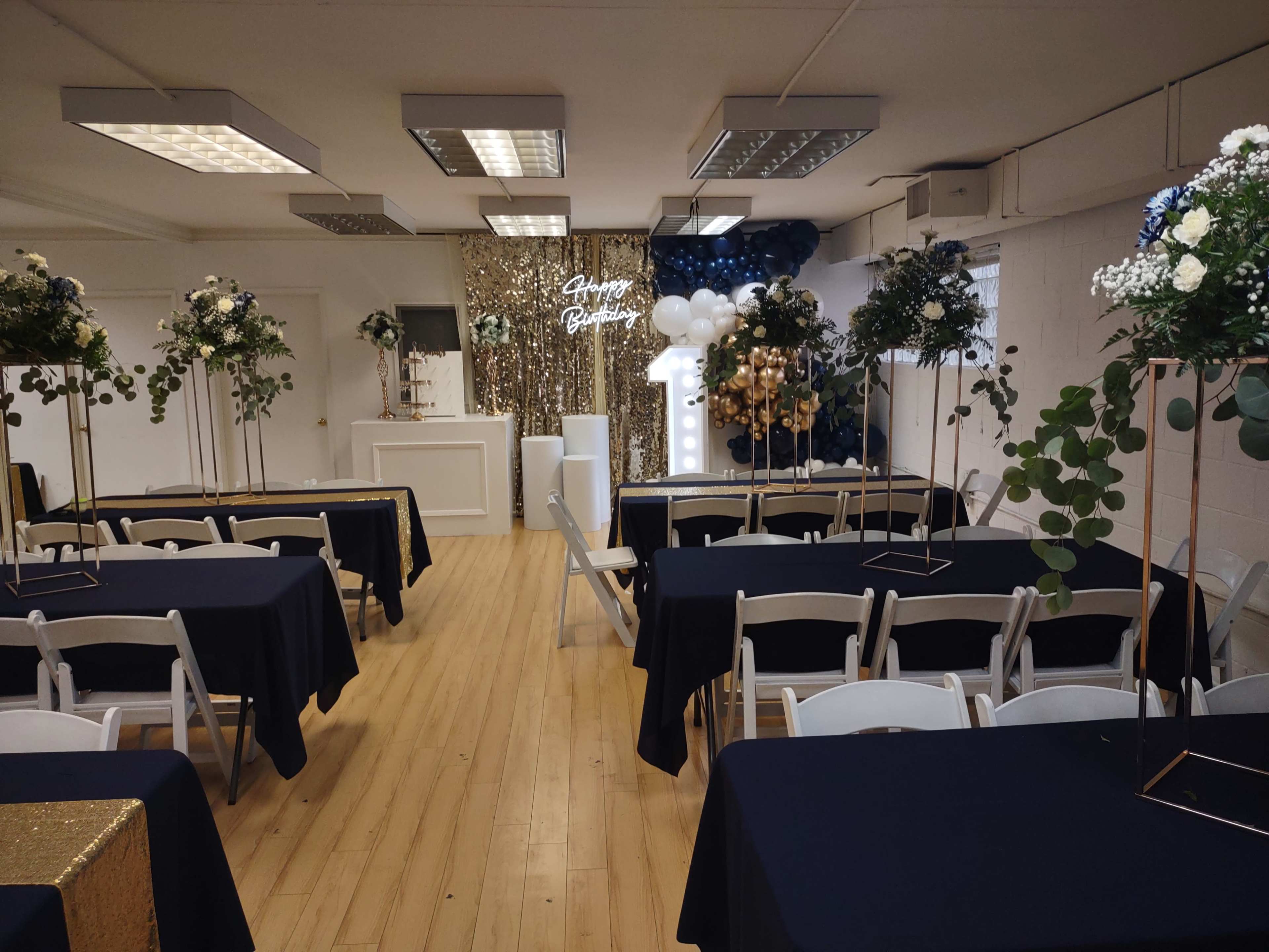 A decorated event space features tables covered with navy cloths, surrounded by greenery, with a backdrop of balloons and a sparkling golden curtain.