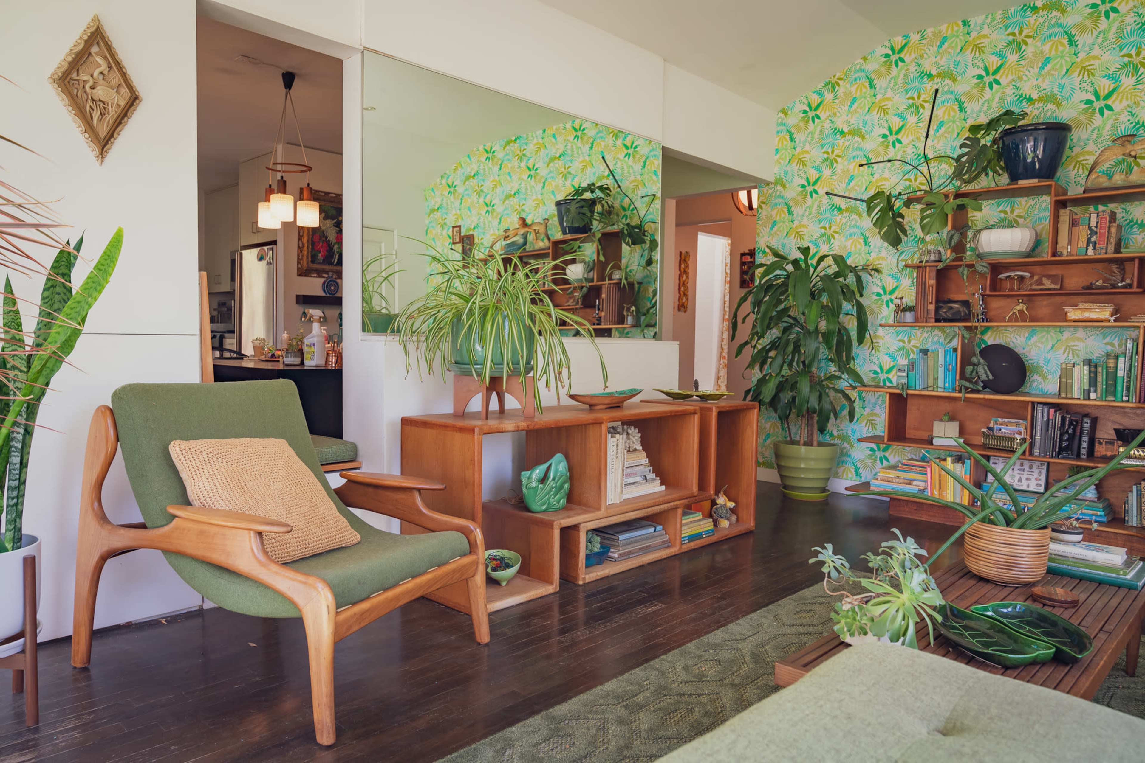 A mid-century modern living room features a green armchair, vibrant tropical wallpaper, and shelves filled with plants and decorative items.