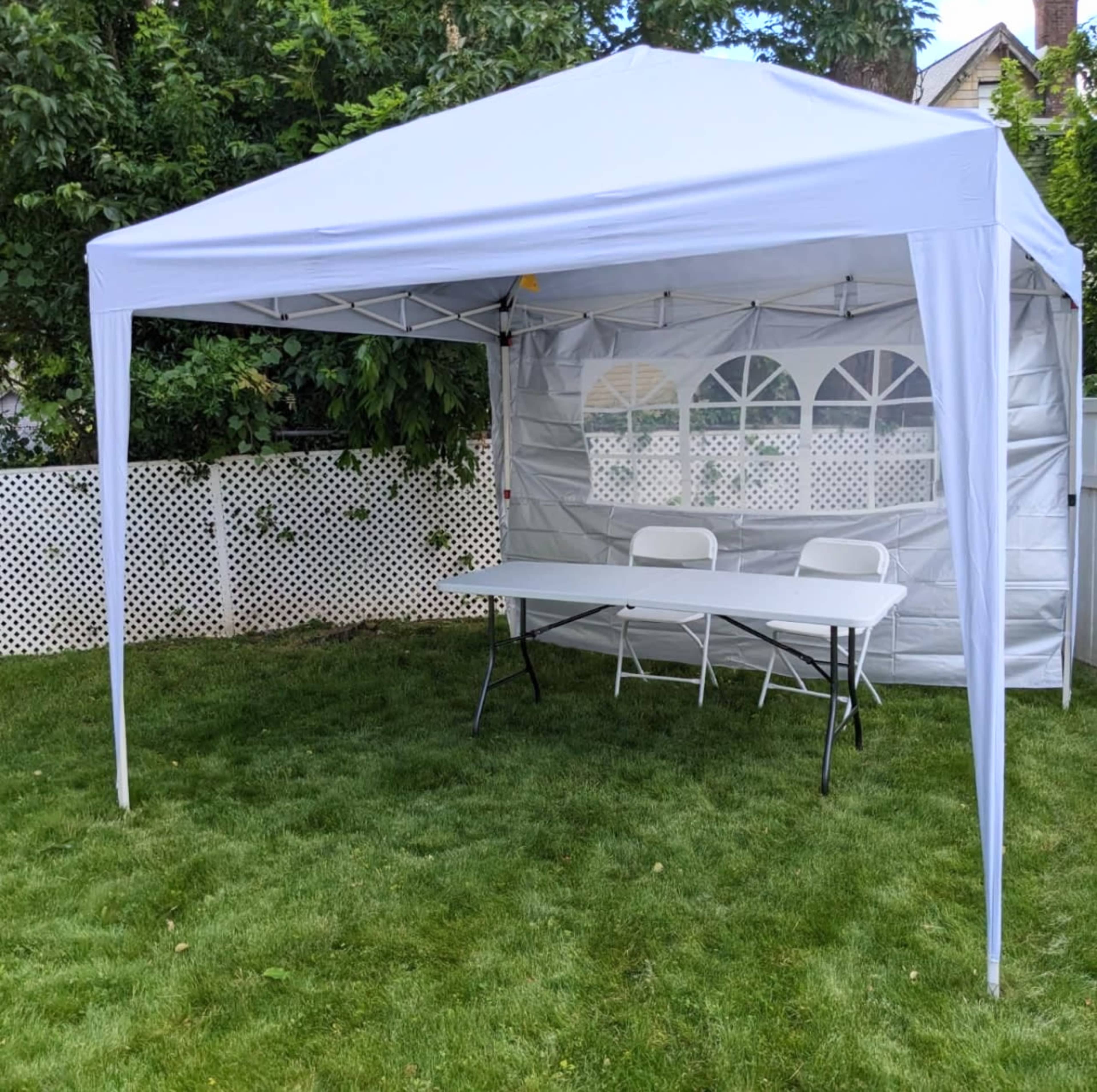 A white canopy tent is set up on green grass with a table and two folding chairs inside, surrounded by a white lattice fence and trees.