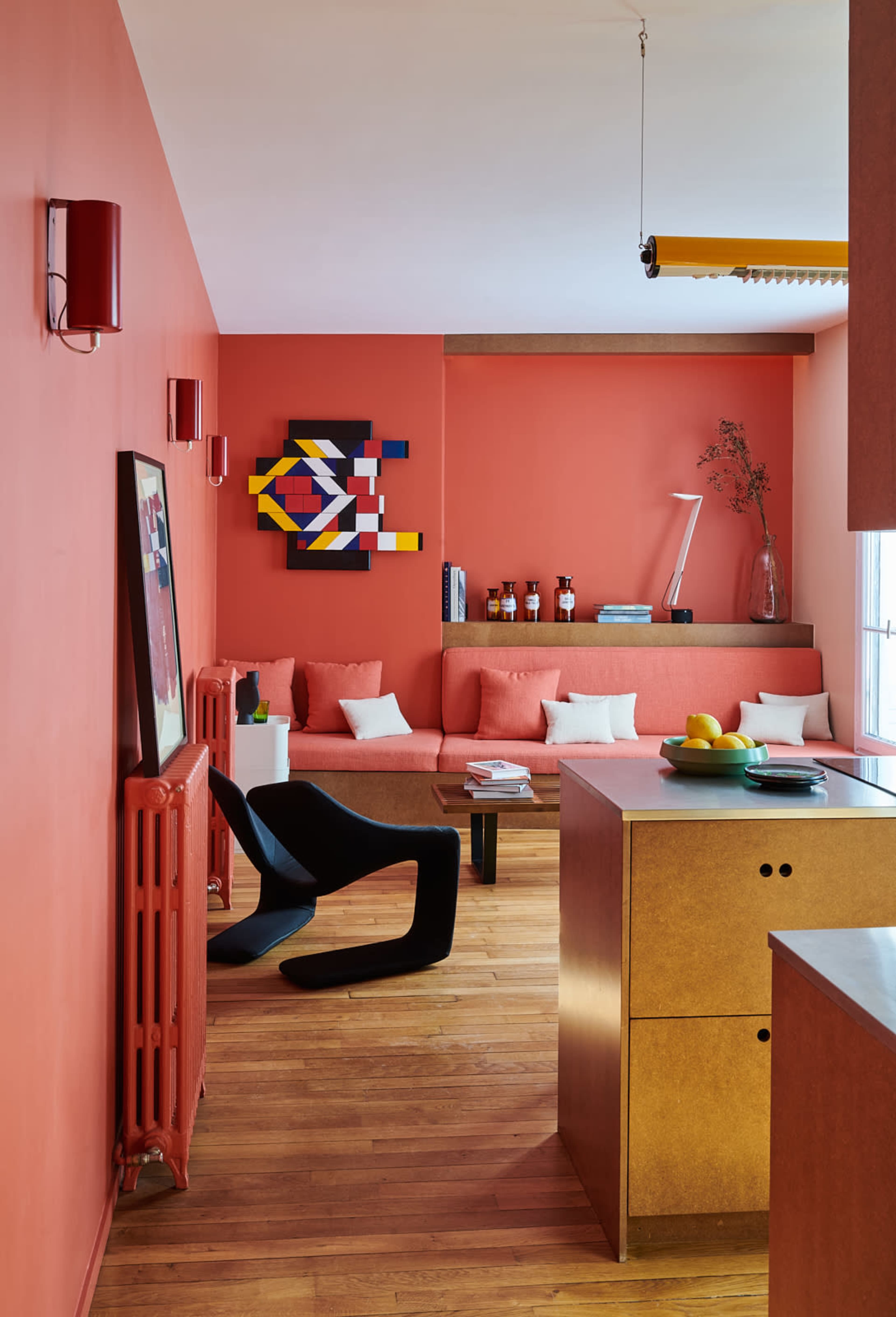 A modern living space with a coral-colored wall, a sectional sofa, and a wooden kitchen island, featuring geometric artwork and decor items.