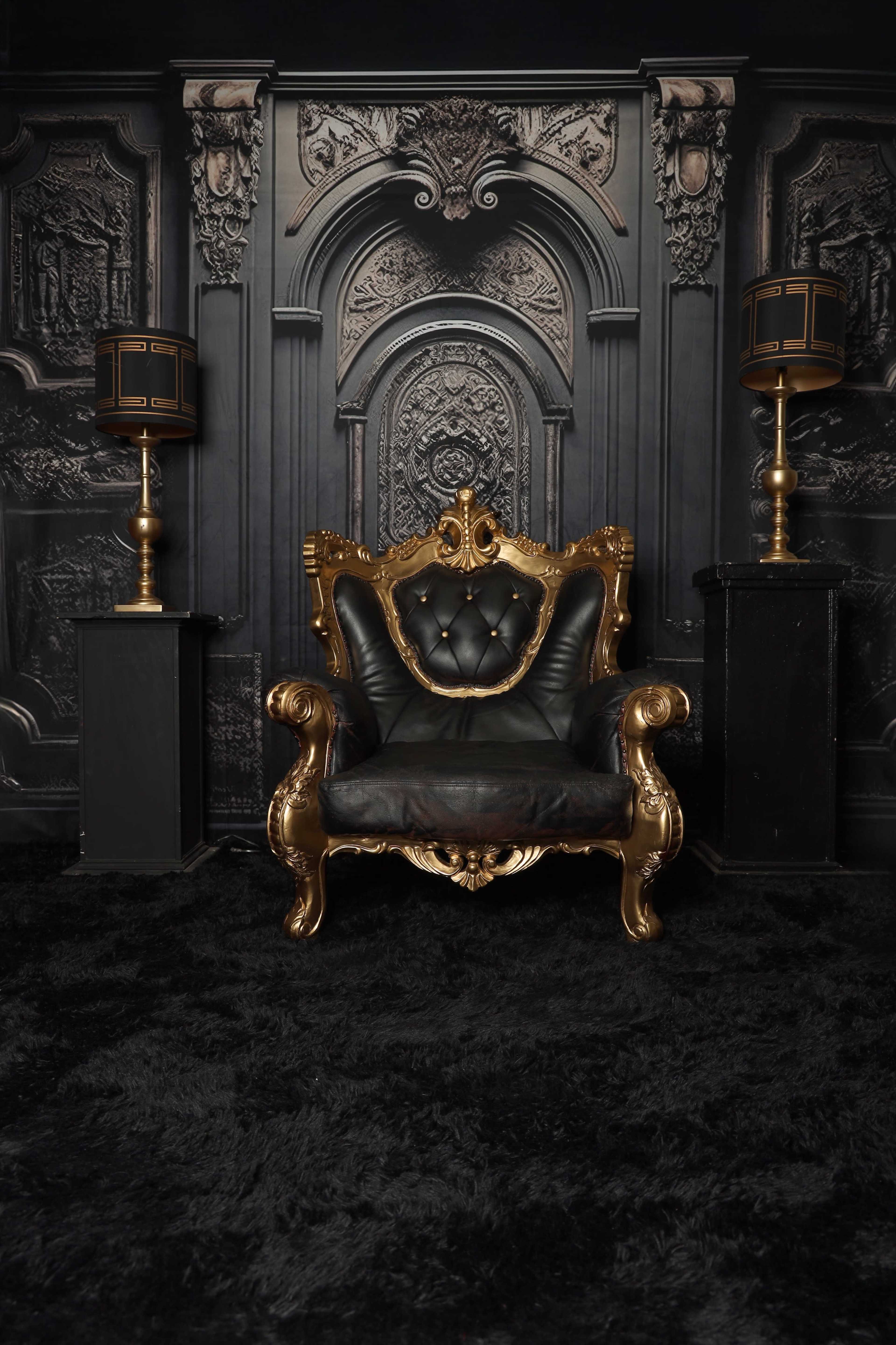 The image shows a luxurious black and gold ornate chair positioned in front of a detailed dark textured wall, with two matching lamps on either side.