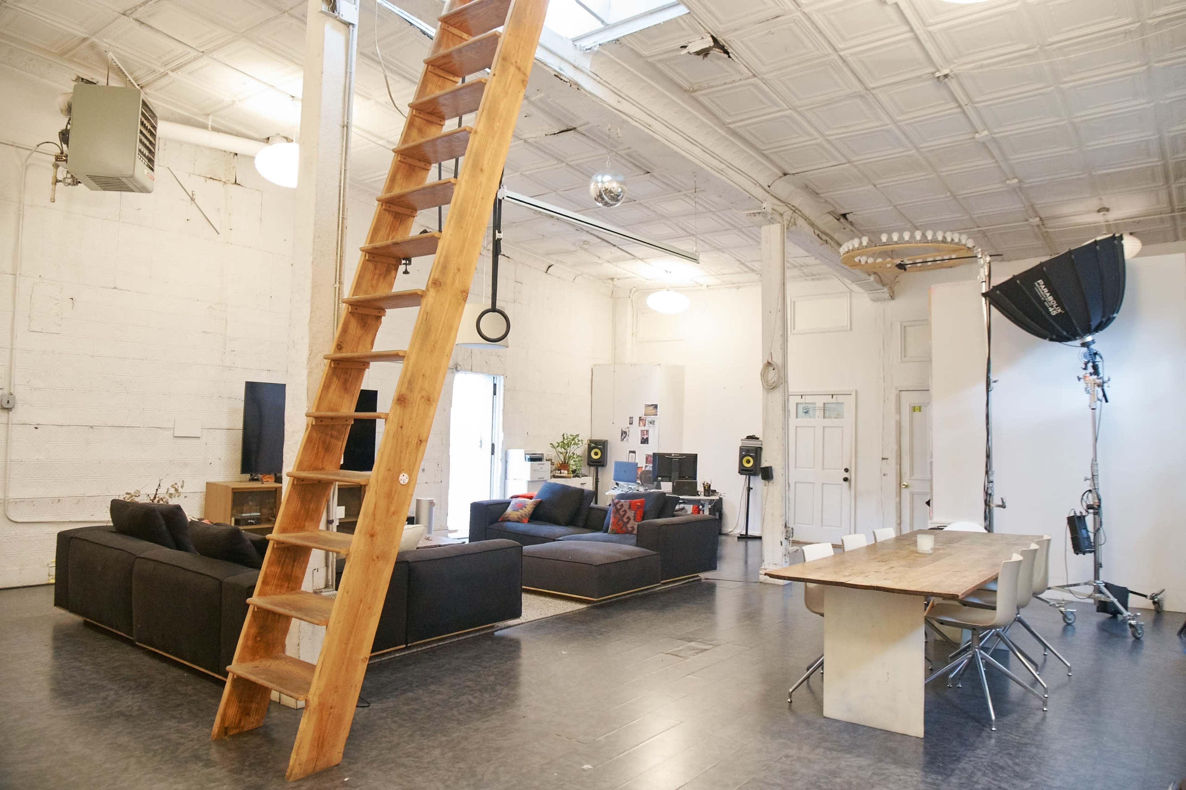 The image shows an open, modern studio space featuring a seating area with dark couches, a wooden ladder leading to a loft, and a large dining table surrounded by chairs.