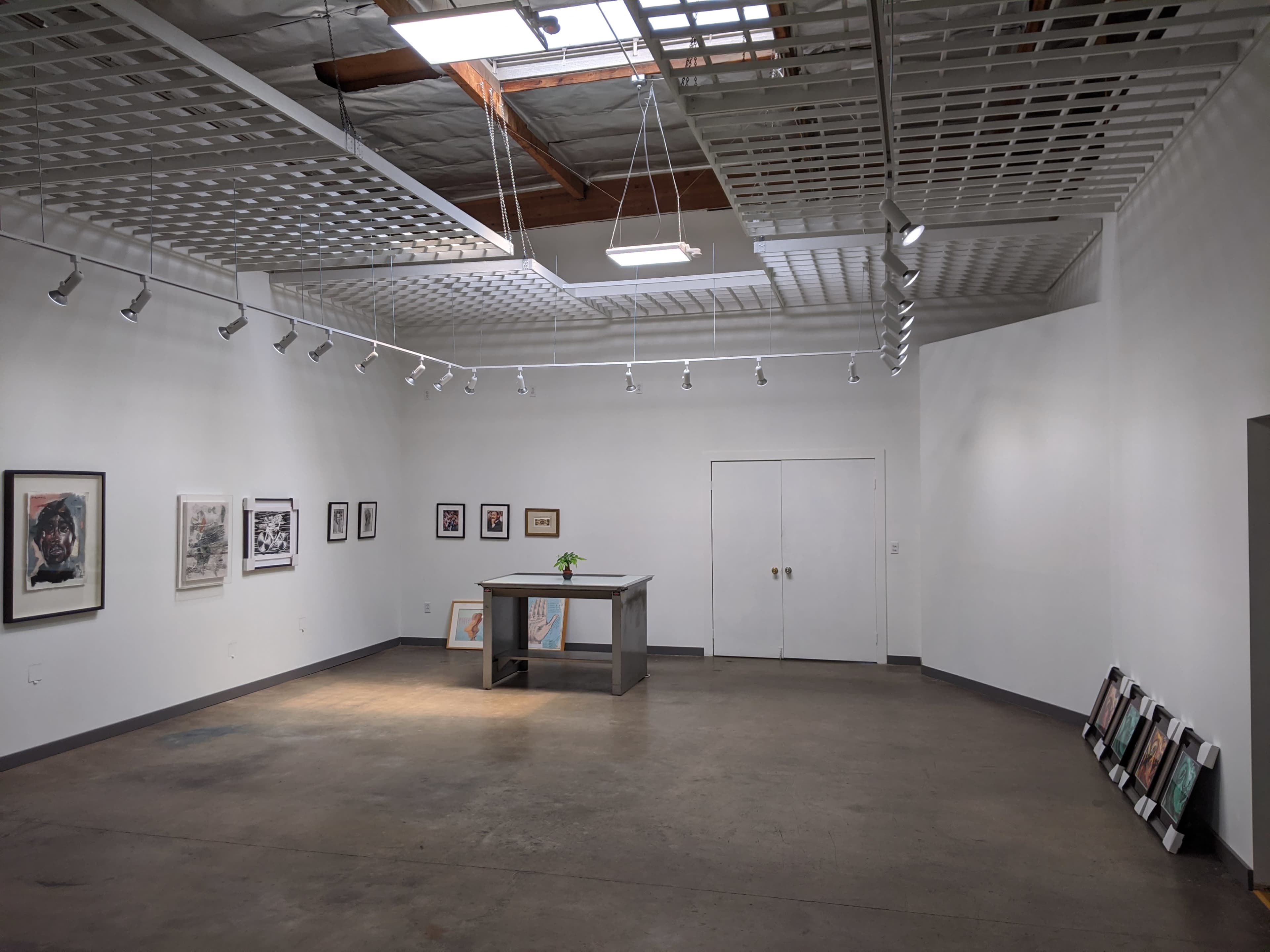 An art gallery features white walls, a central gray display table with a small plant, and framed artworks along the sides.