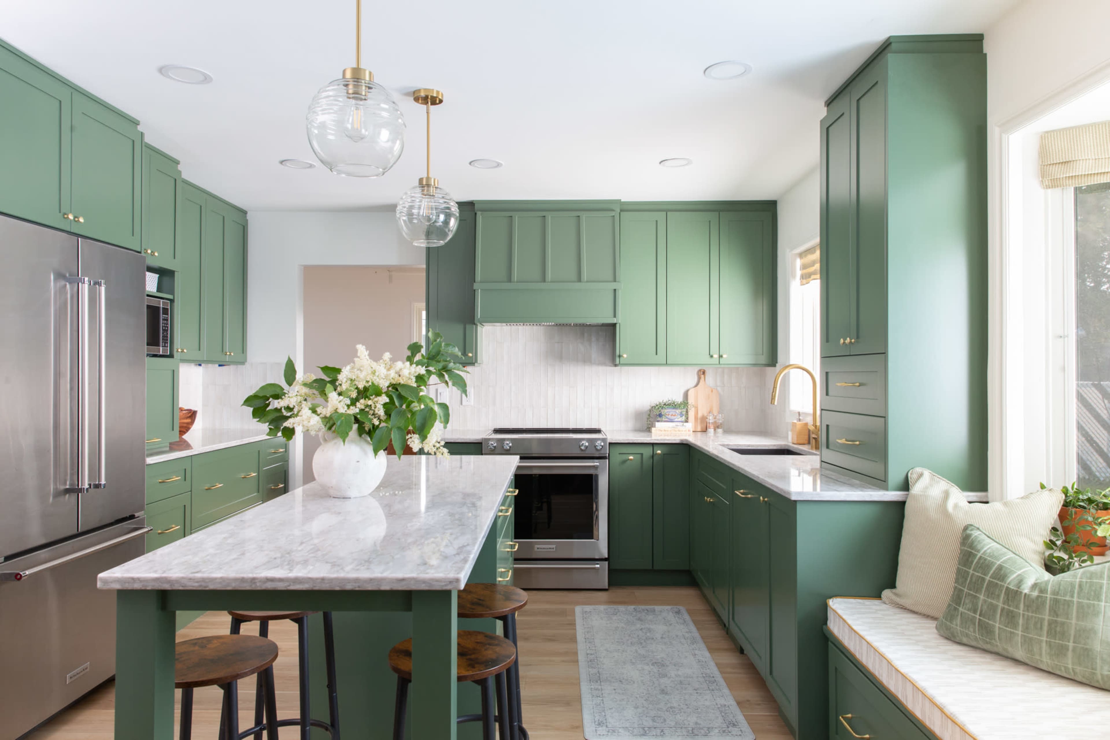 The image shows a modern kitchen with green cabinets, a marble countertop, stainless steel appliances, and a cozy bench seating area.