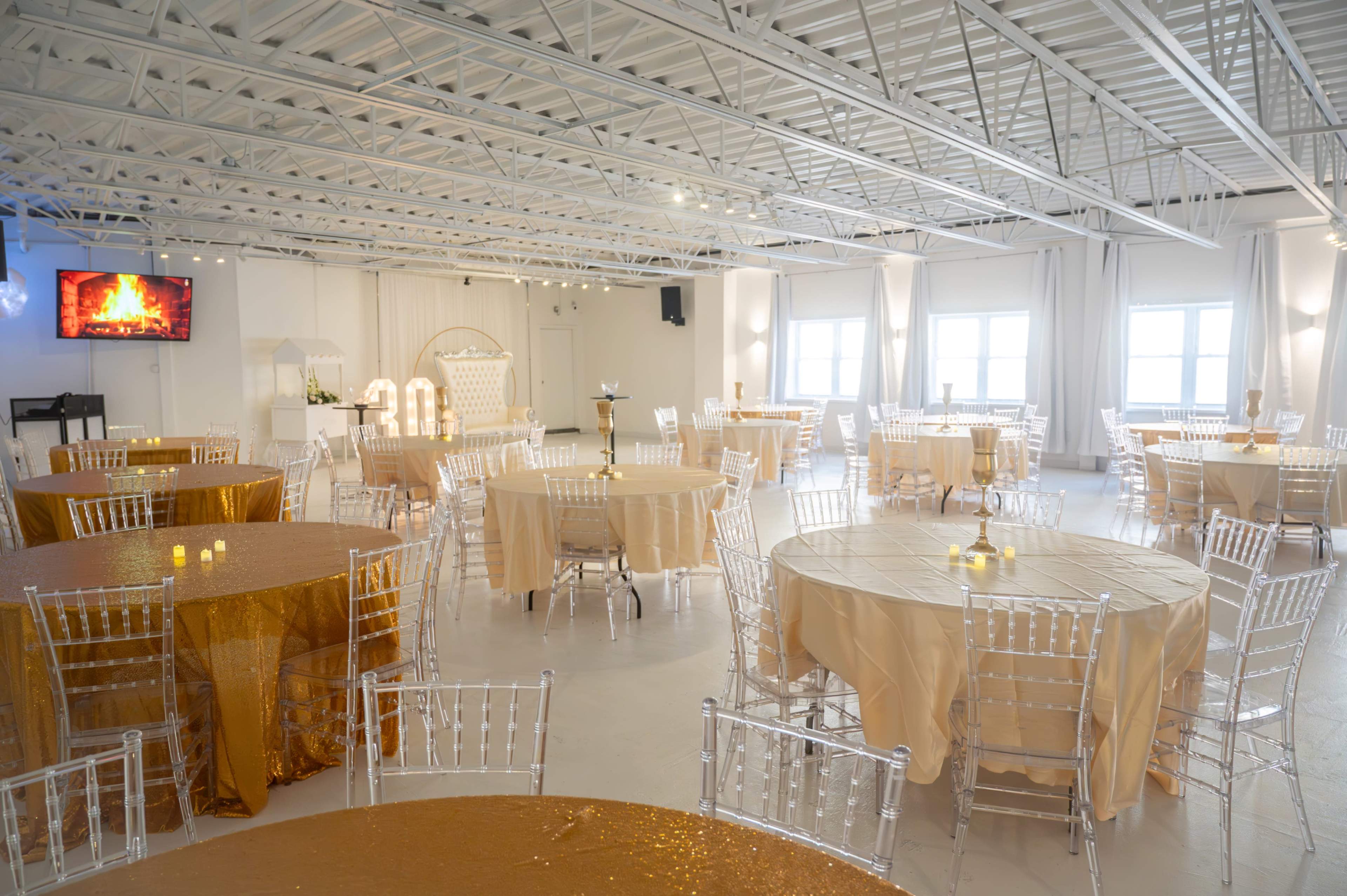 A spacious event room features multiple round tables set with golden tablecloths and transparent chiavari chairs, with a large screen displaying a flame in the background.