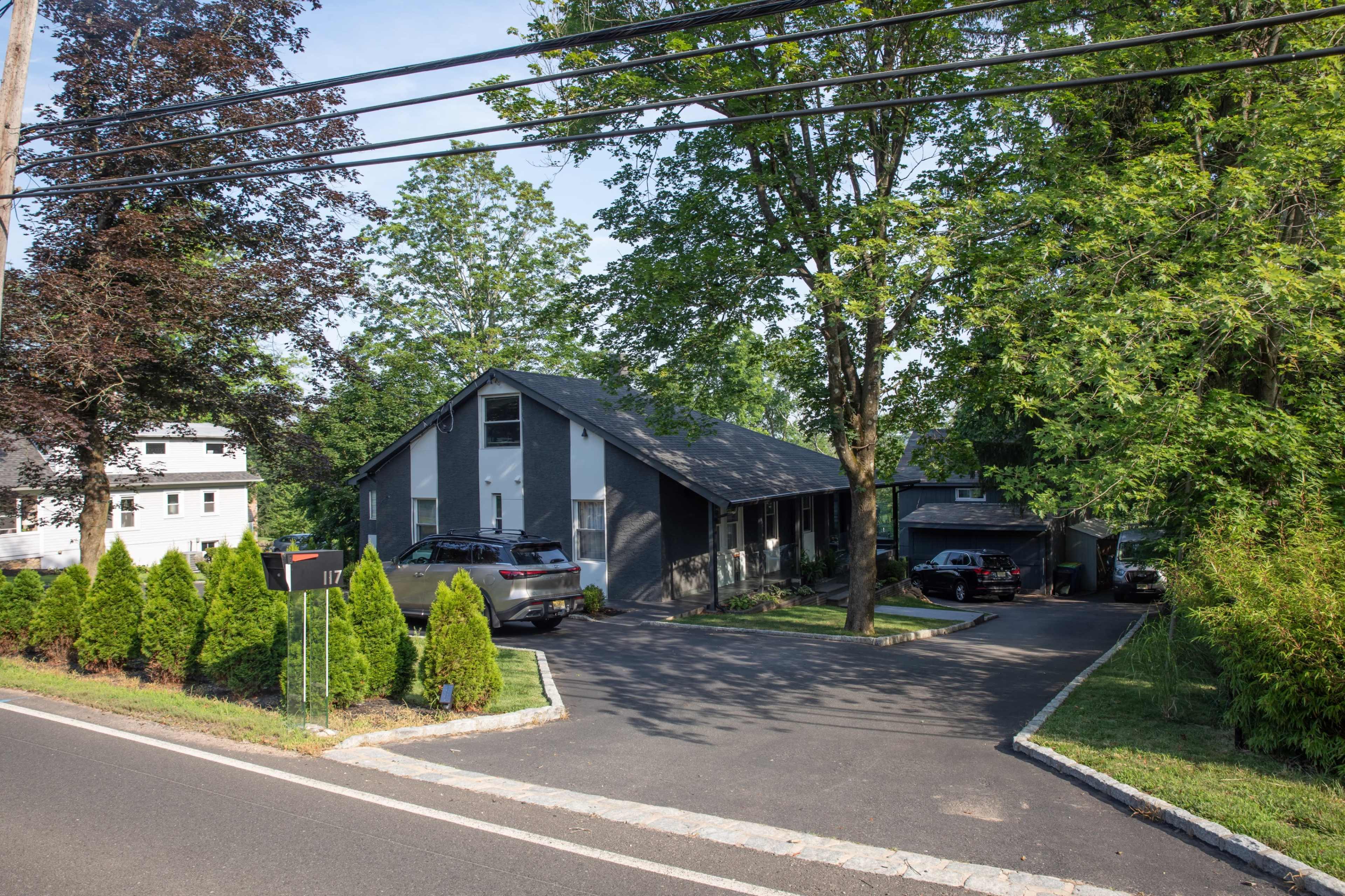 A gray house with a sloped roof is surrounded by trees and has a driveway with parked cars.
