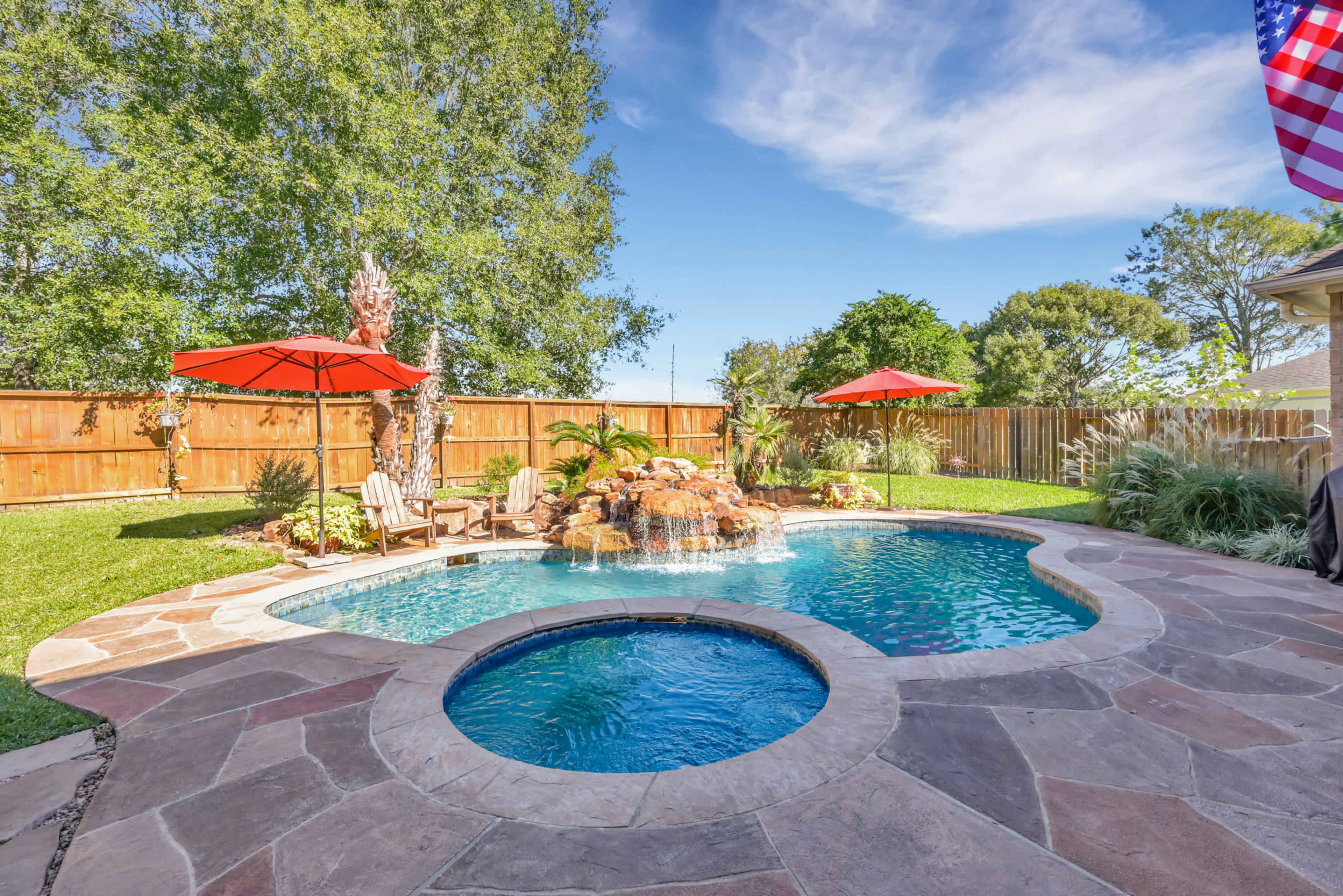 A backyard with a swimming pool featuring a rock waterfall, surrounded by chairs, umbrellas, and landscaped greenery.
