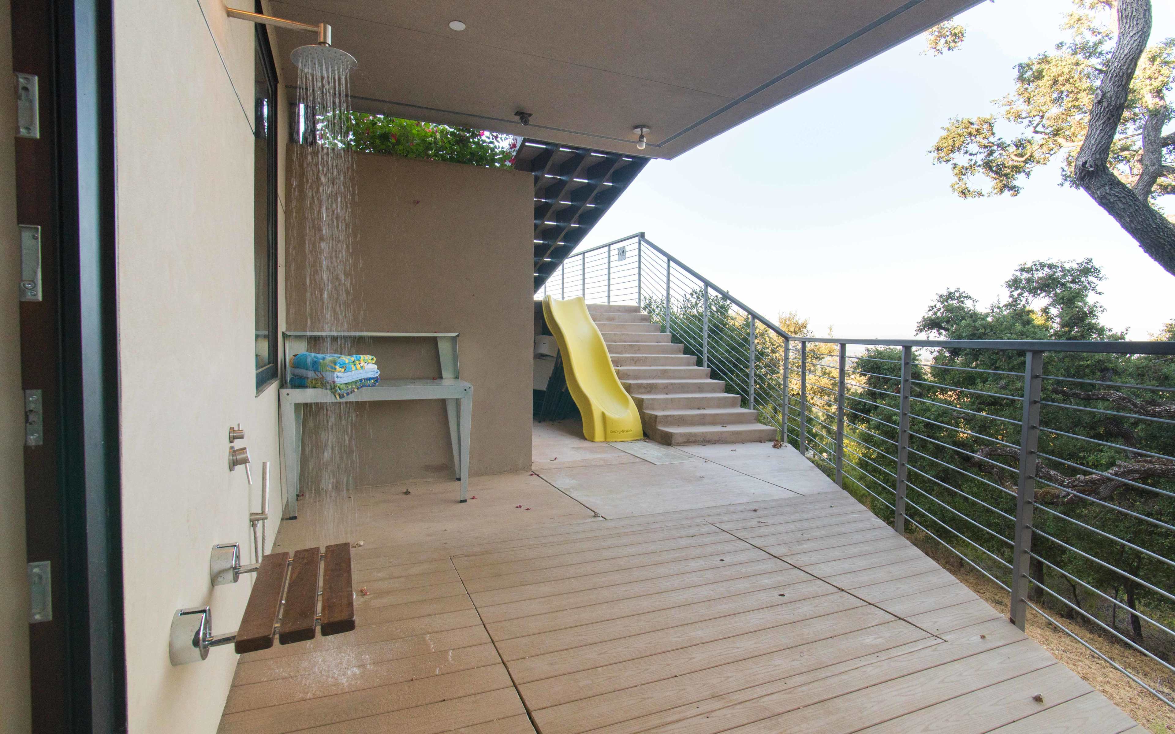 A patio area with a rain shower, a wooden bench, a slide, and stairs leading up to a deck.
