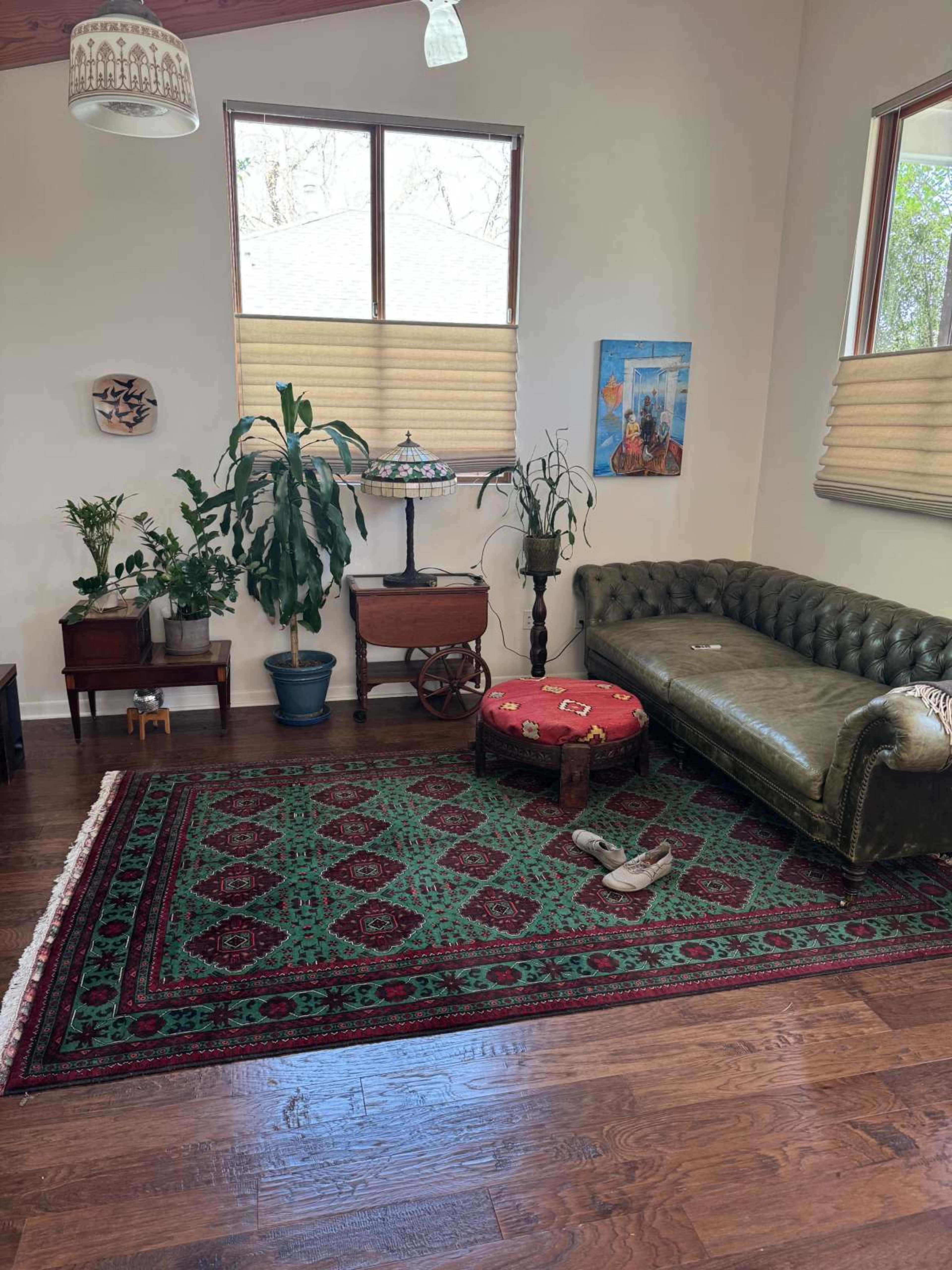 The image shows a cozy living room featuring a green tufted sofa, a patterned rug, potted plants, and a small wooden table with a decorative piece.