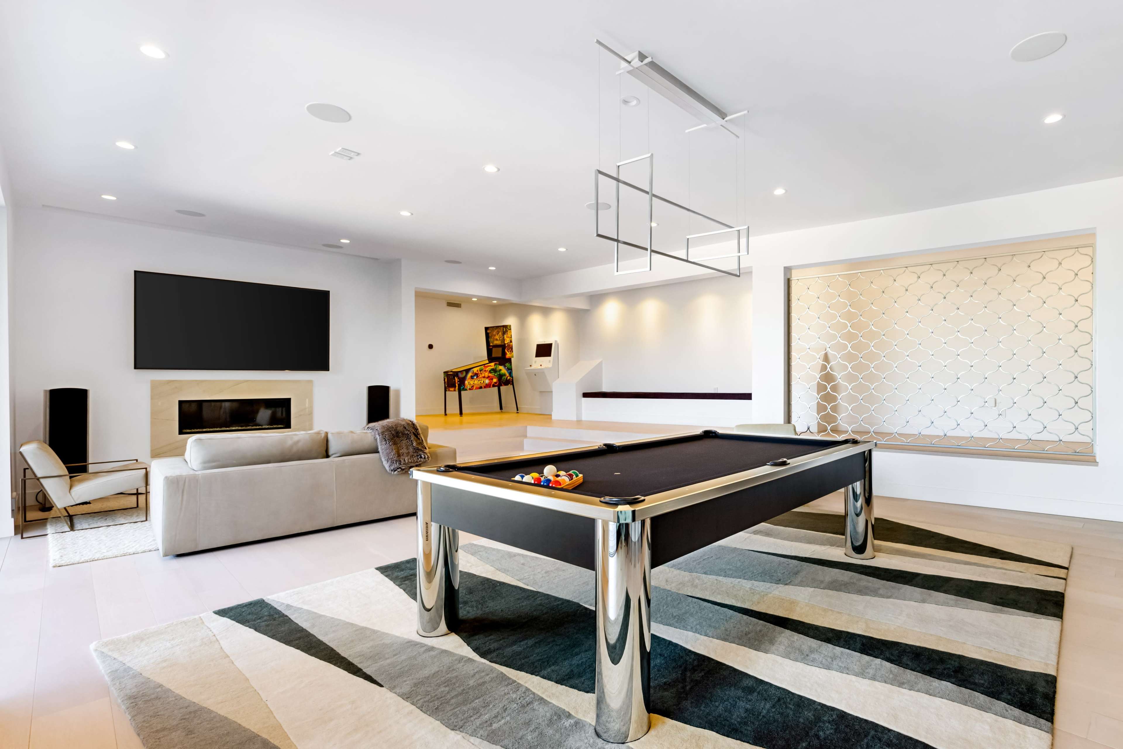 The image shows a modern, spacious game room featuring a pool table, a large television mounted on the wall, a cozy sofa, and an artistic wall design.