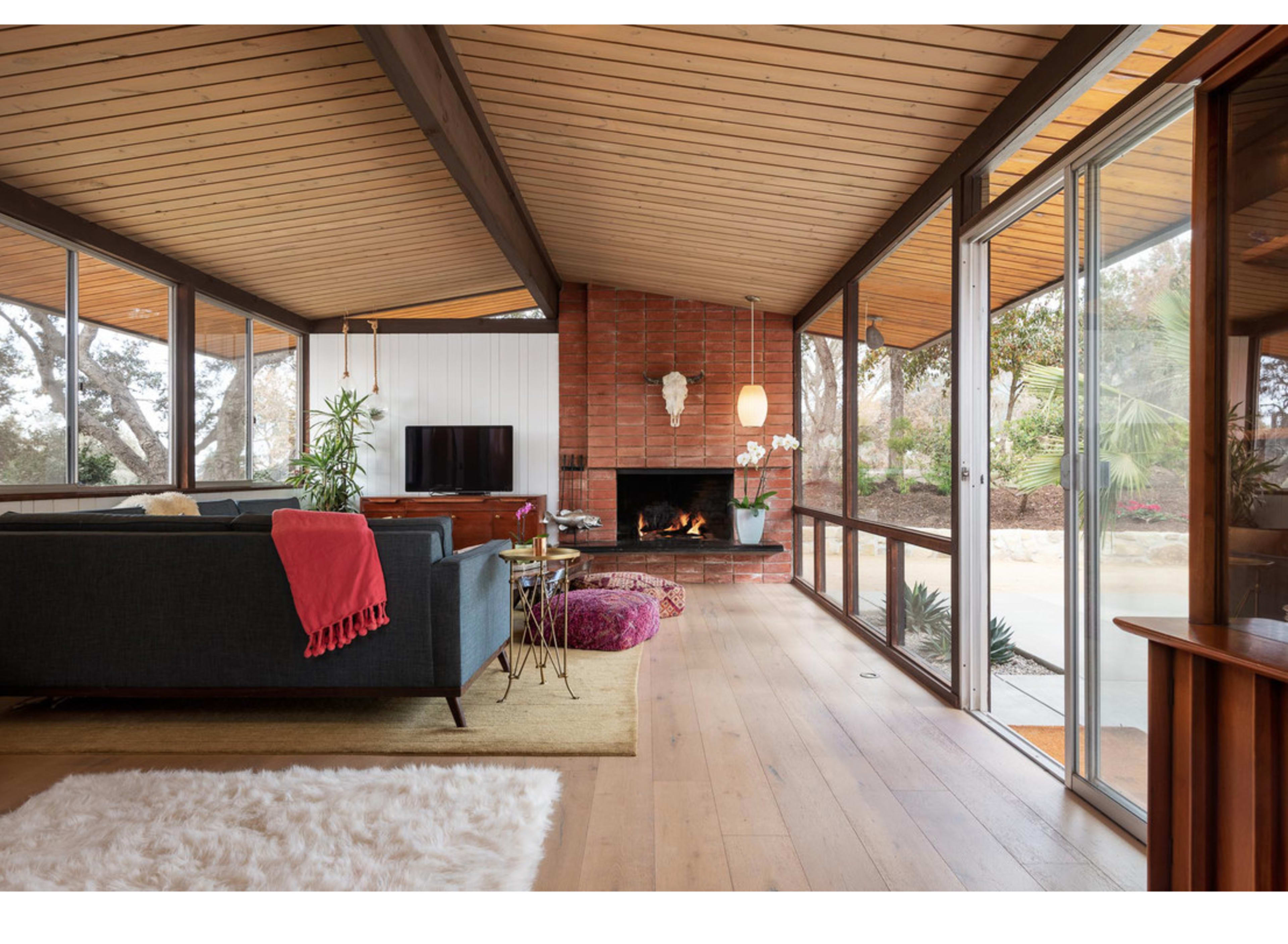 The image shows a modern living room with large windows, wooden ceilings, and a fireplace surrounded by a carpet, furniture, and decorative plants.