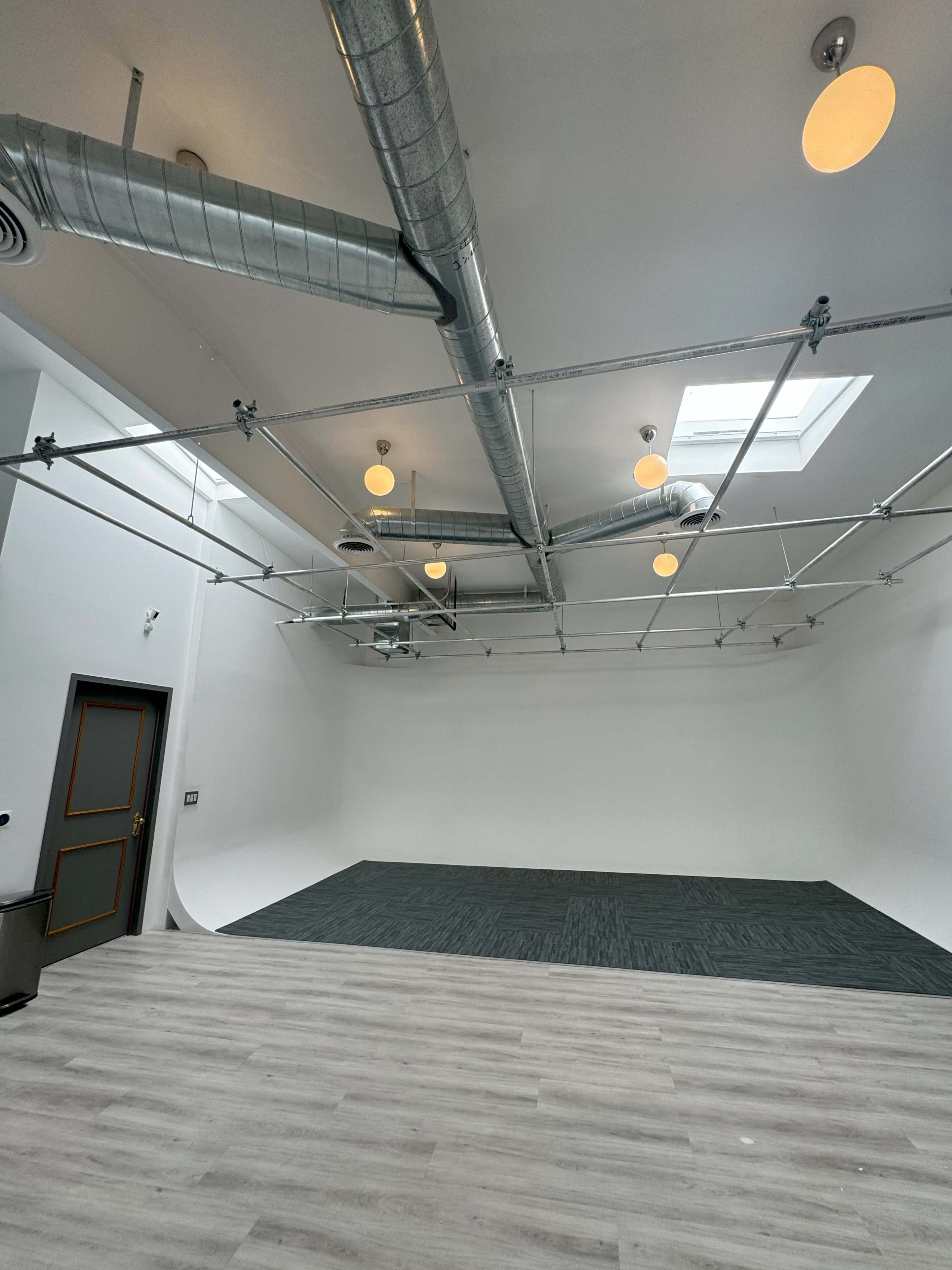 A spacious, empty studio with a white backdrop, gray flooring, and visible ceiling ducts and lights.