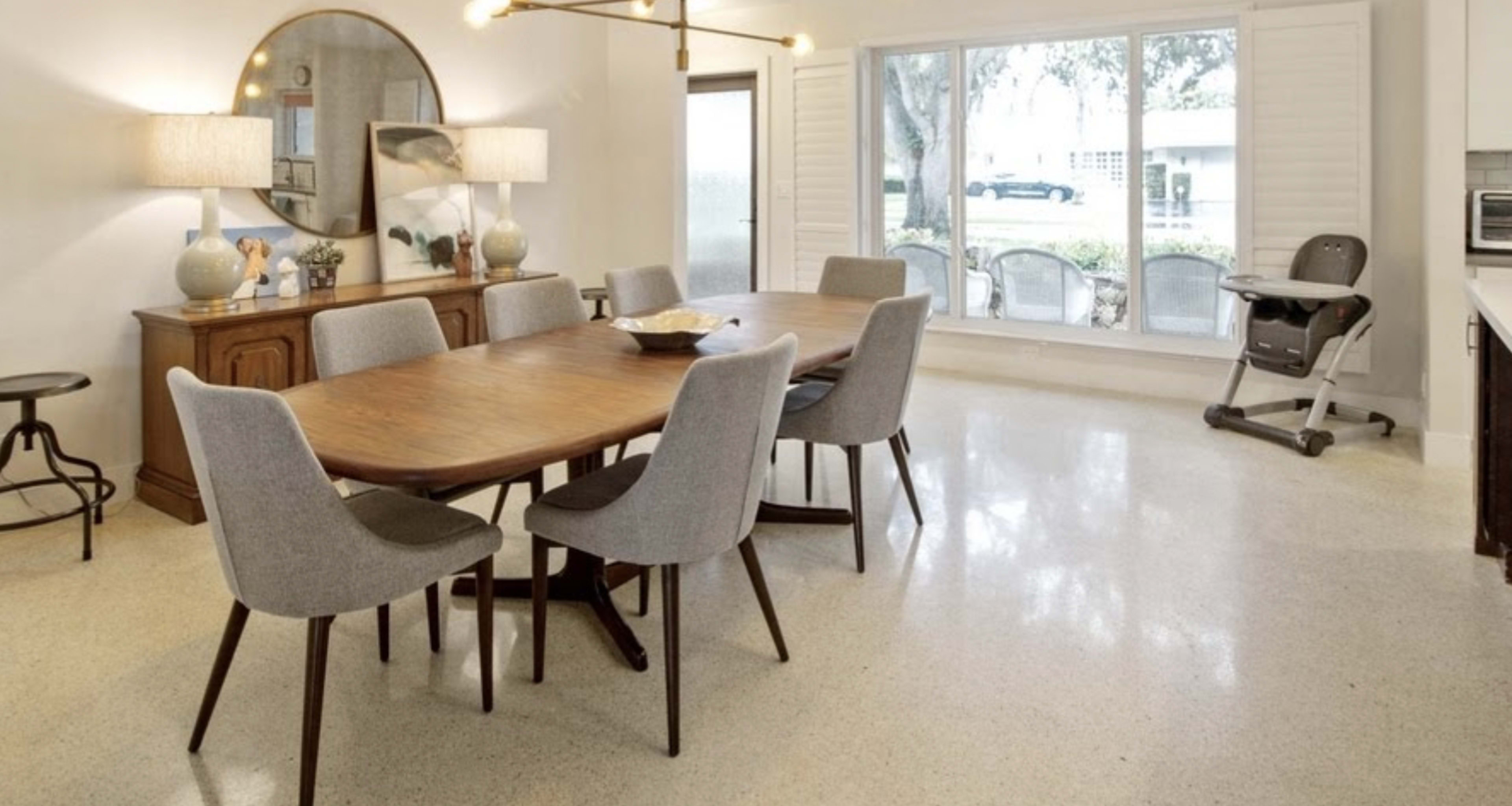 A dining area features a wooden table surrounded by six gray chairs, with a sideboard and a large mirror on the wall, and a high chair positioned near a window.