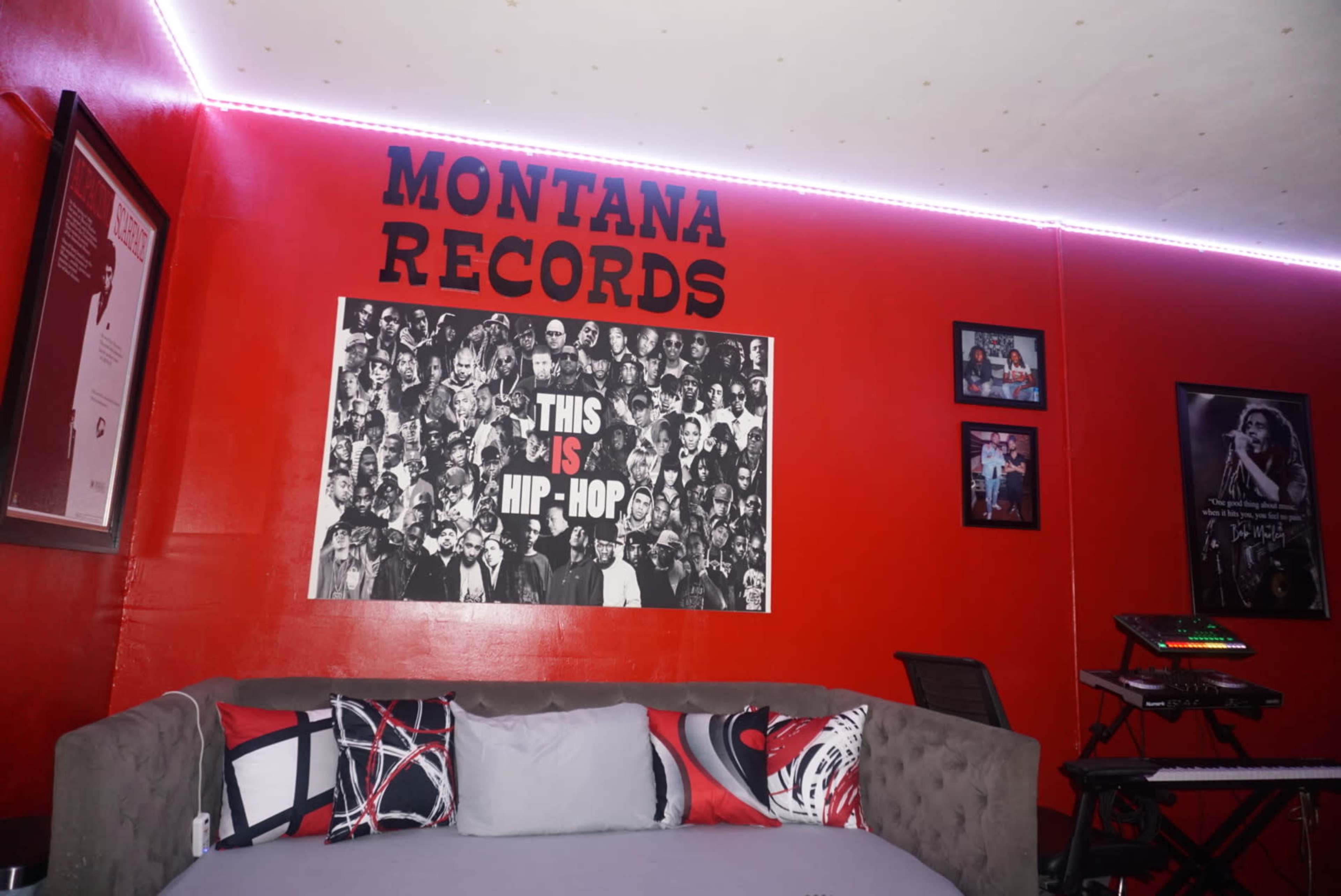 The image shows a room with a red wall featuring the text "MONTANA RECORDS" and a large poster of hip-hop artists, alongside framed pictures and a music setup.