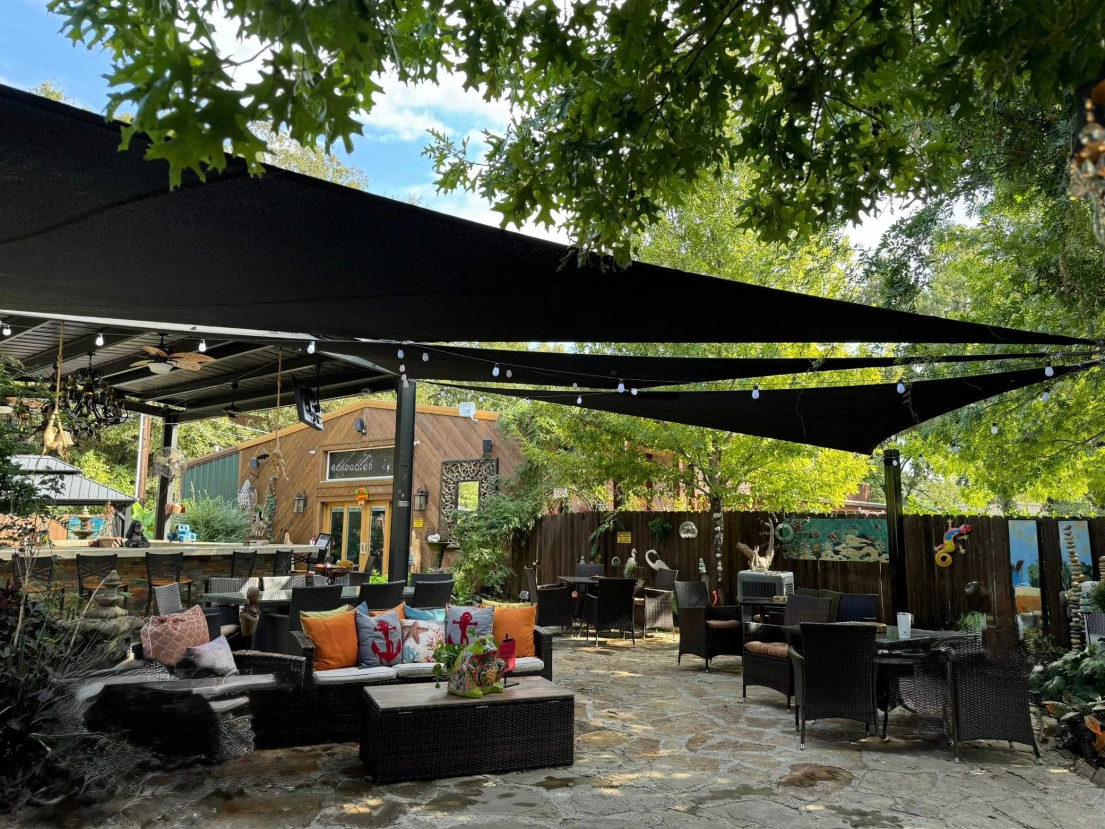 The image shows a shaded outdoor seating area with cushioned furniture, decorative plants, and an inviting atmosphere under a canopy of fabric and string lights.