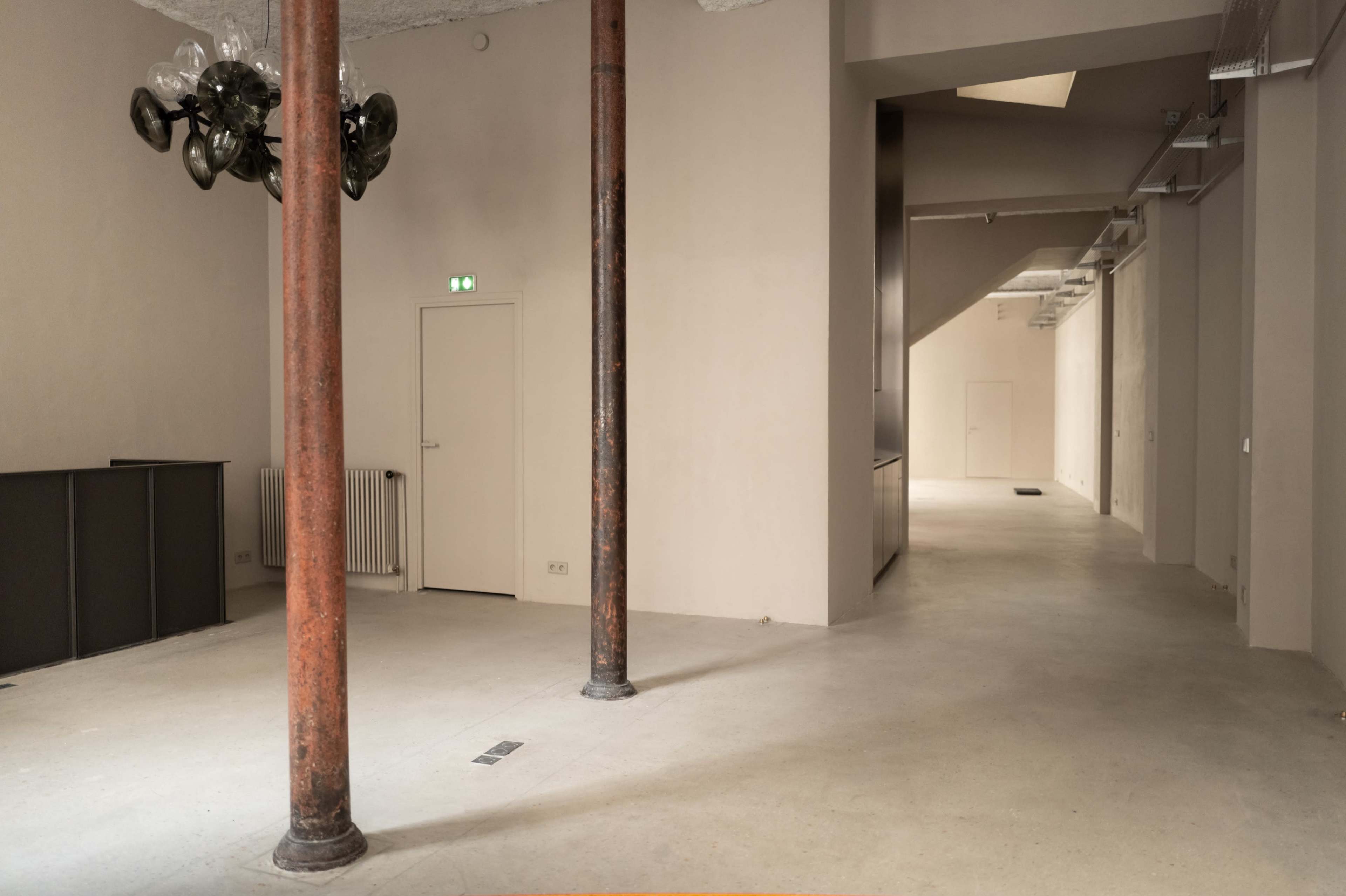 The image shows a large, empty interior space with columns, a door on one side, and a hallway leading to another area.