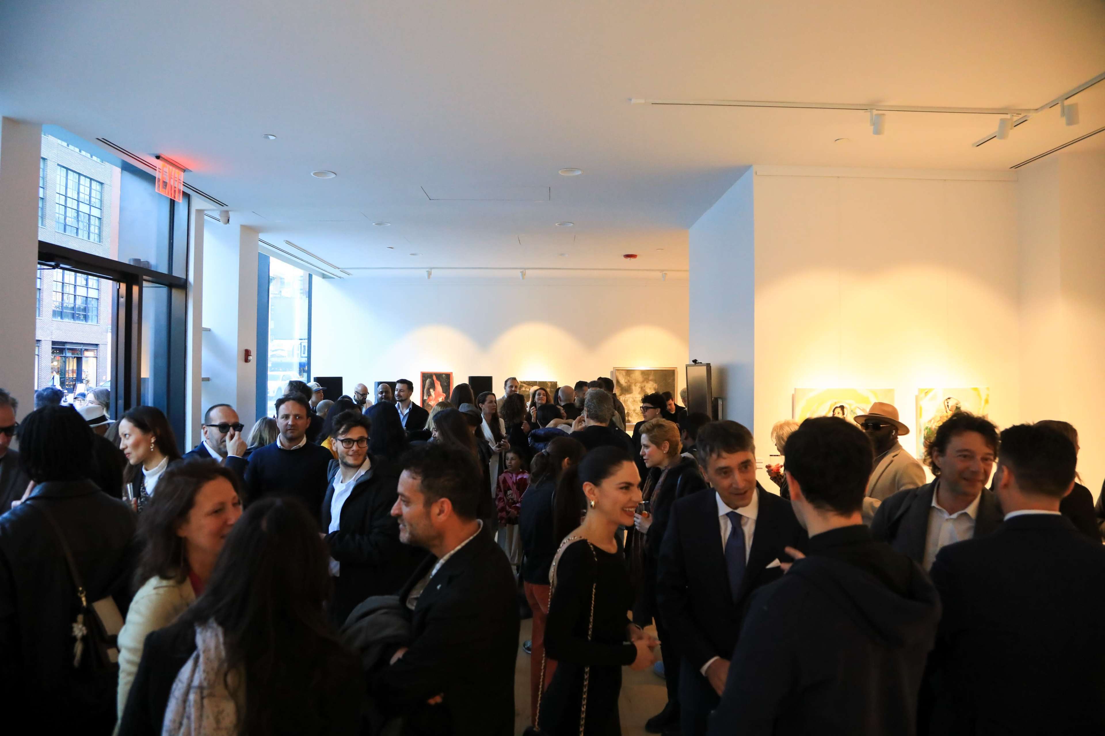 A large group of people mingles in an art gallery with white walls and large windows overlooking the street.