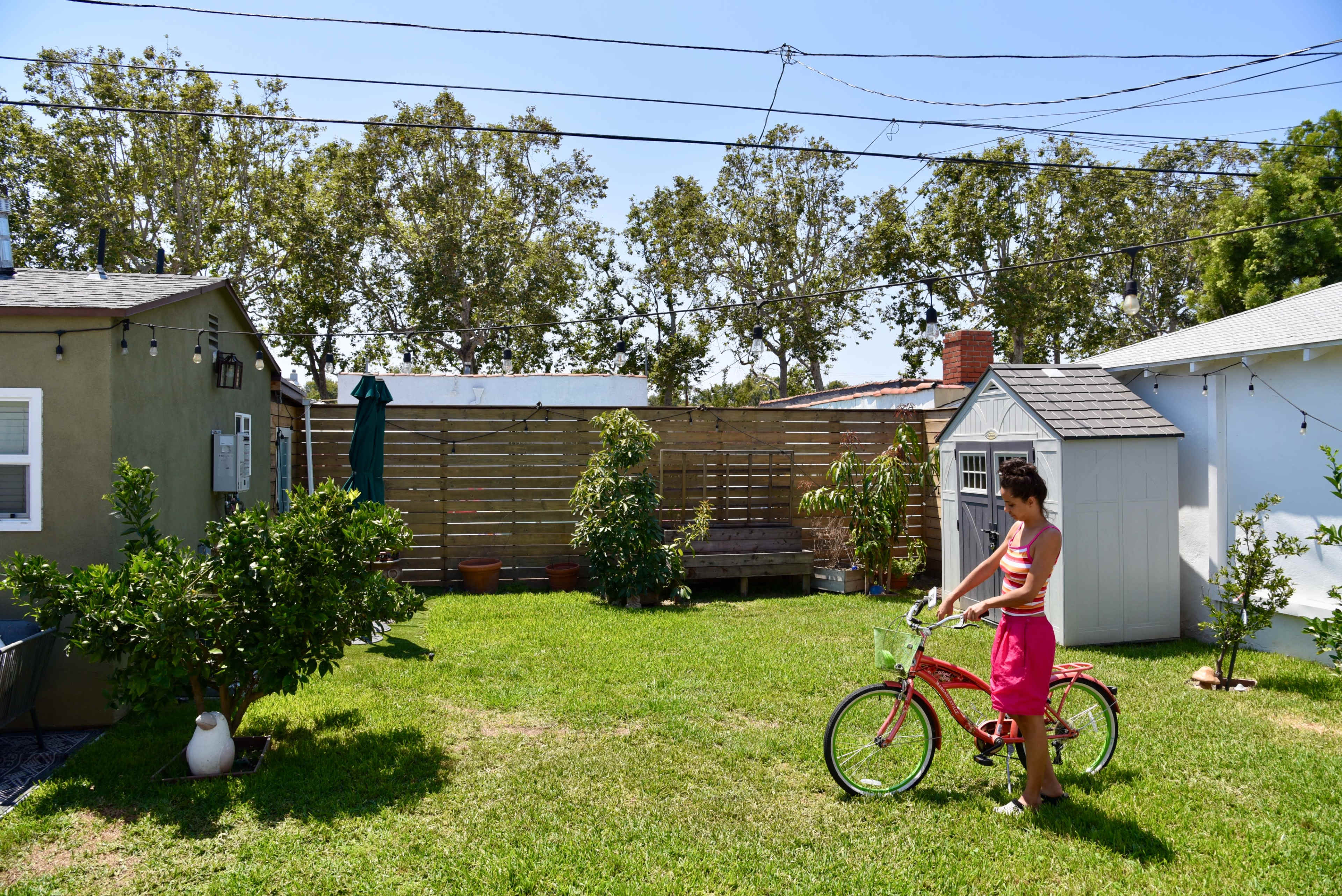 A child stands beside a pink bicycle in a grassy backyard surrounded by small structures and trees.