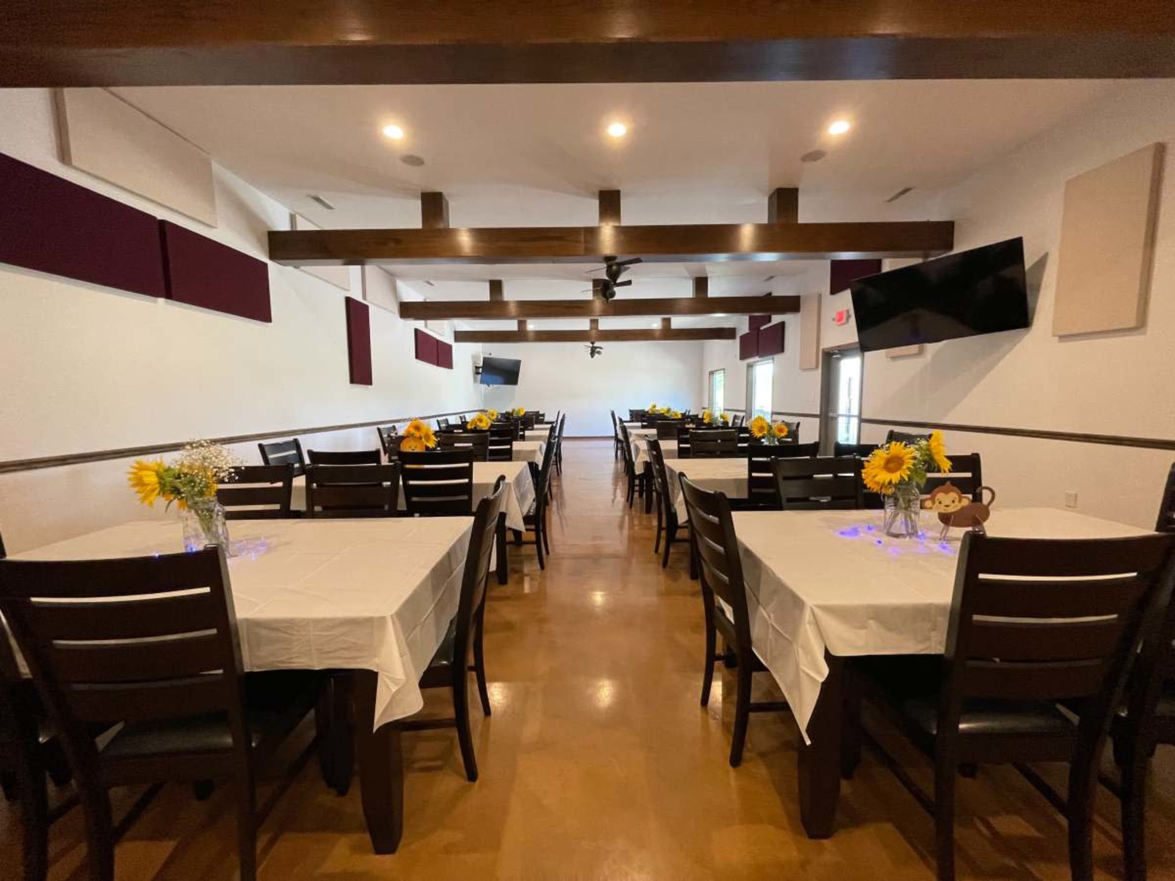 A large, well-lit room with multiple rows of tables covered in white tablecloths, each adorned with sunflower centerpieces.
