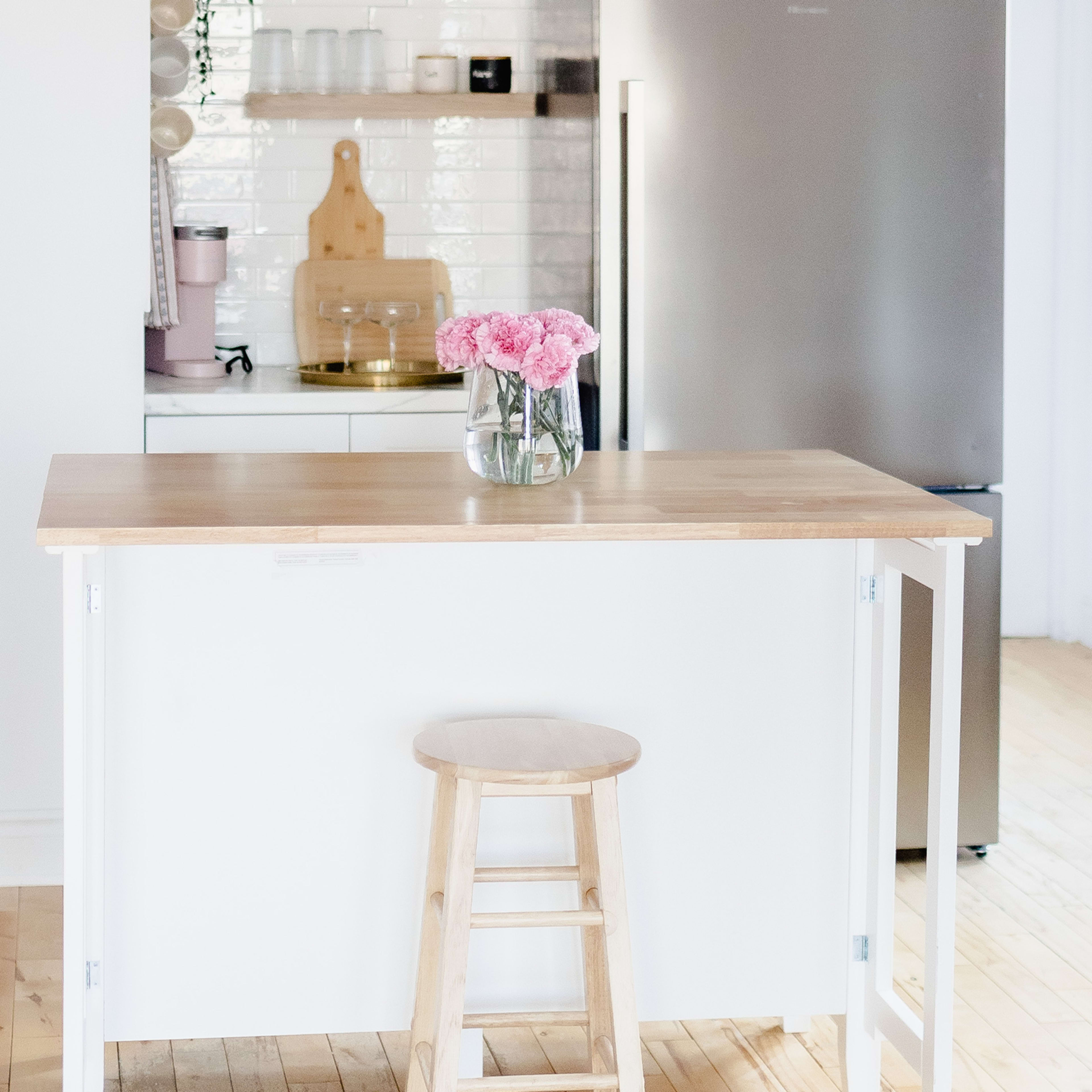 A wooden table with a stool and a vase of pink flowers is positioned in a bright kitchen with white cabinets and a stainless steel refrigerator.