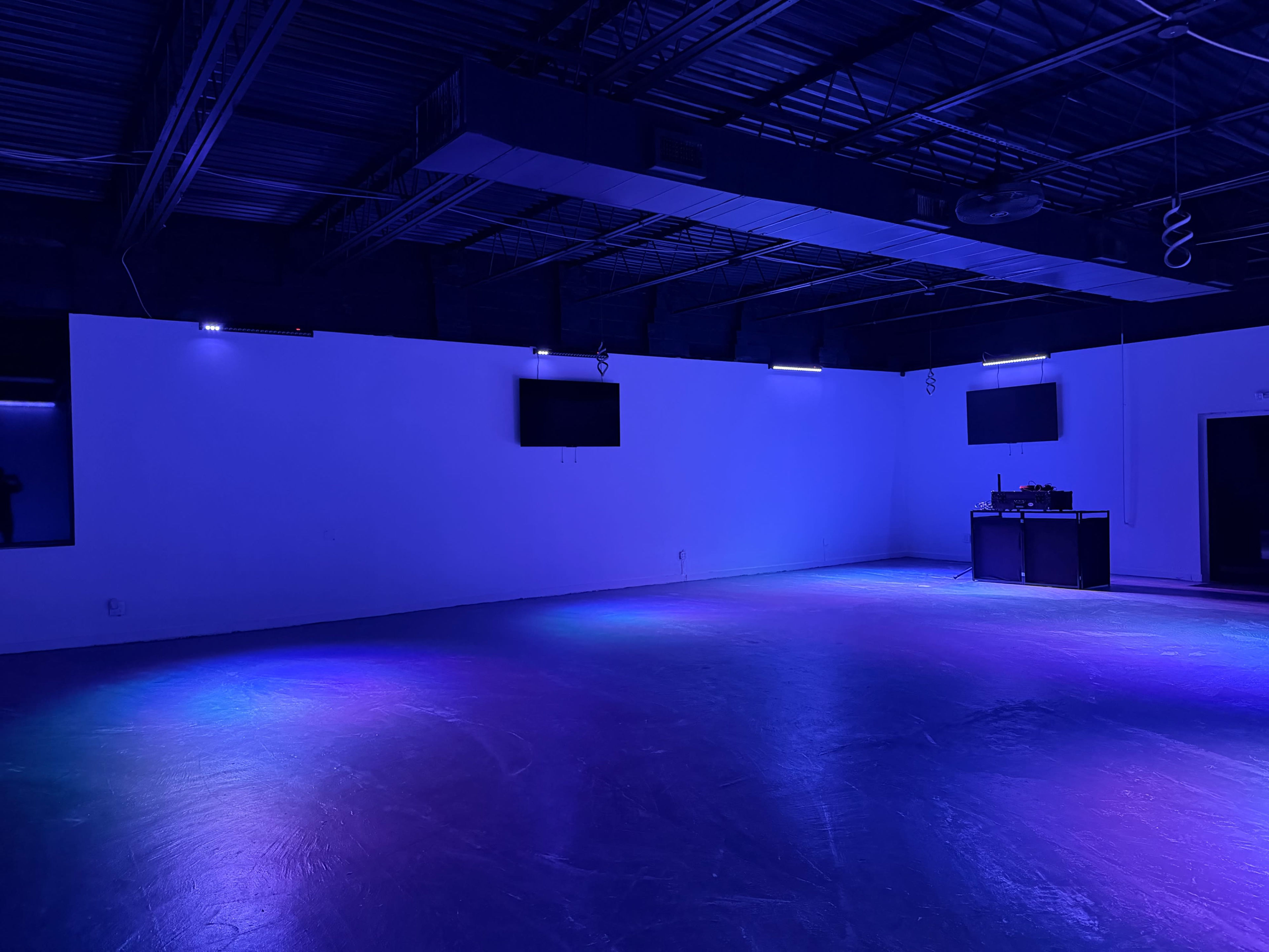 The image shows an empty room with blue and purple lighting, featuring a DJ setup on one side and two wall-mounted screens.