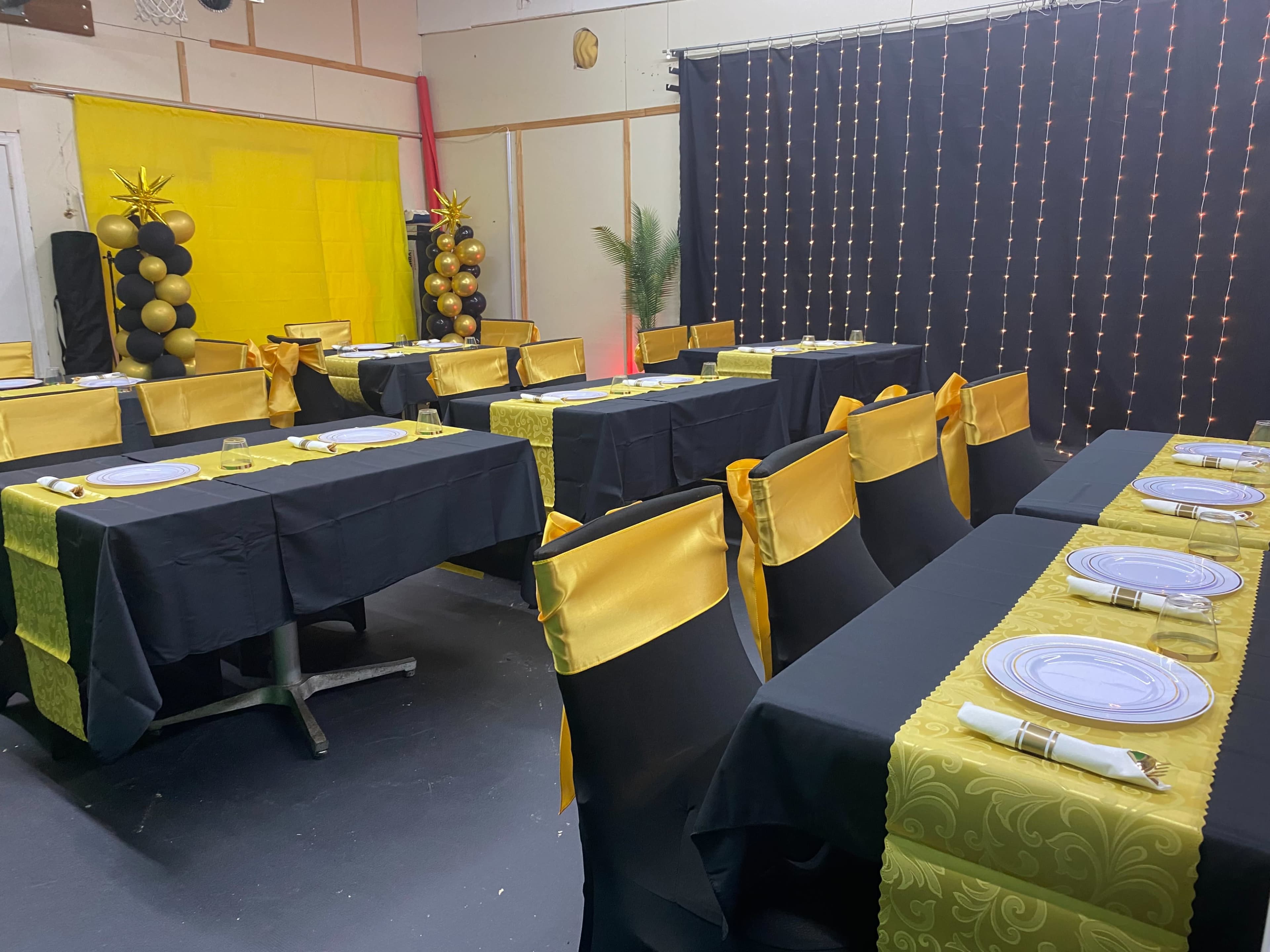 The image shows a decorated event space with several tables covered in black and yellow tablecloths, set with plates and utensils, and surrounded by string lights and balloon columns.