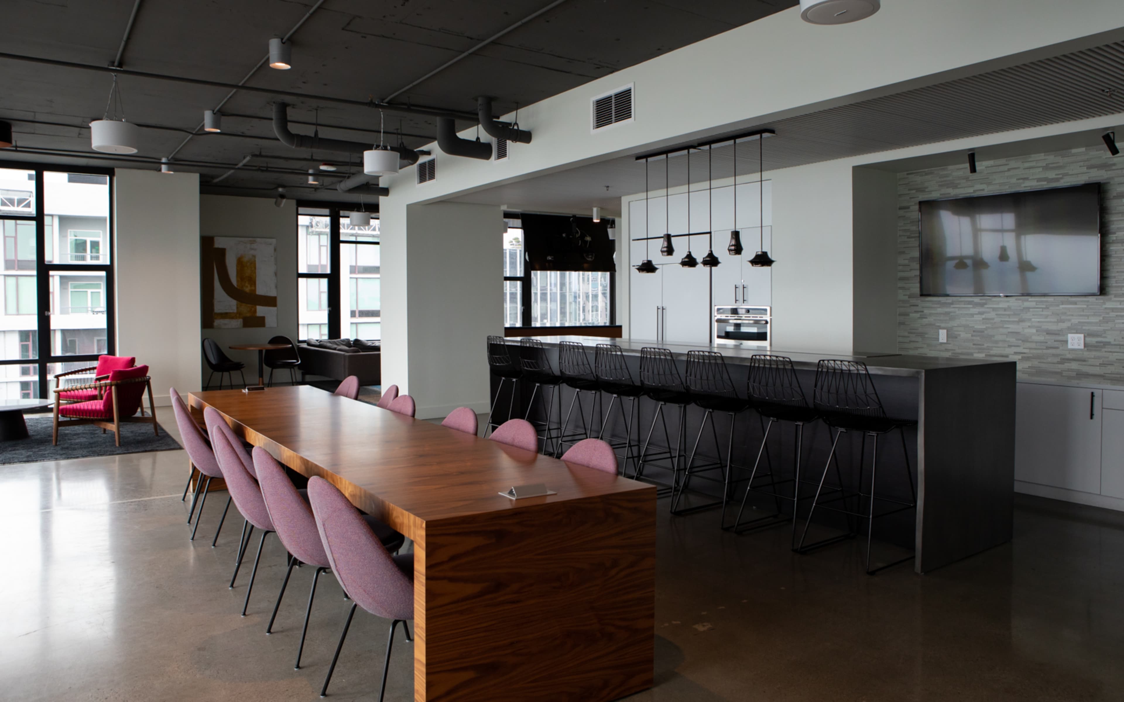 A modern office kitchen area featuring a long wooden table, black bar stools, and large windows with a view.