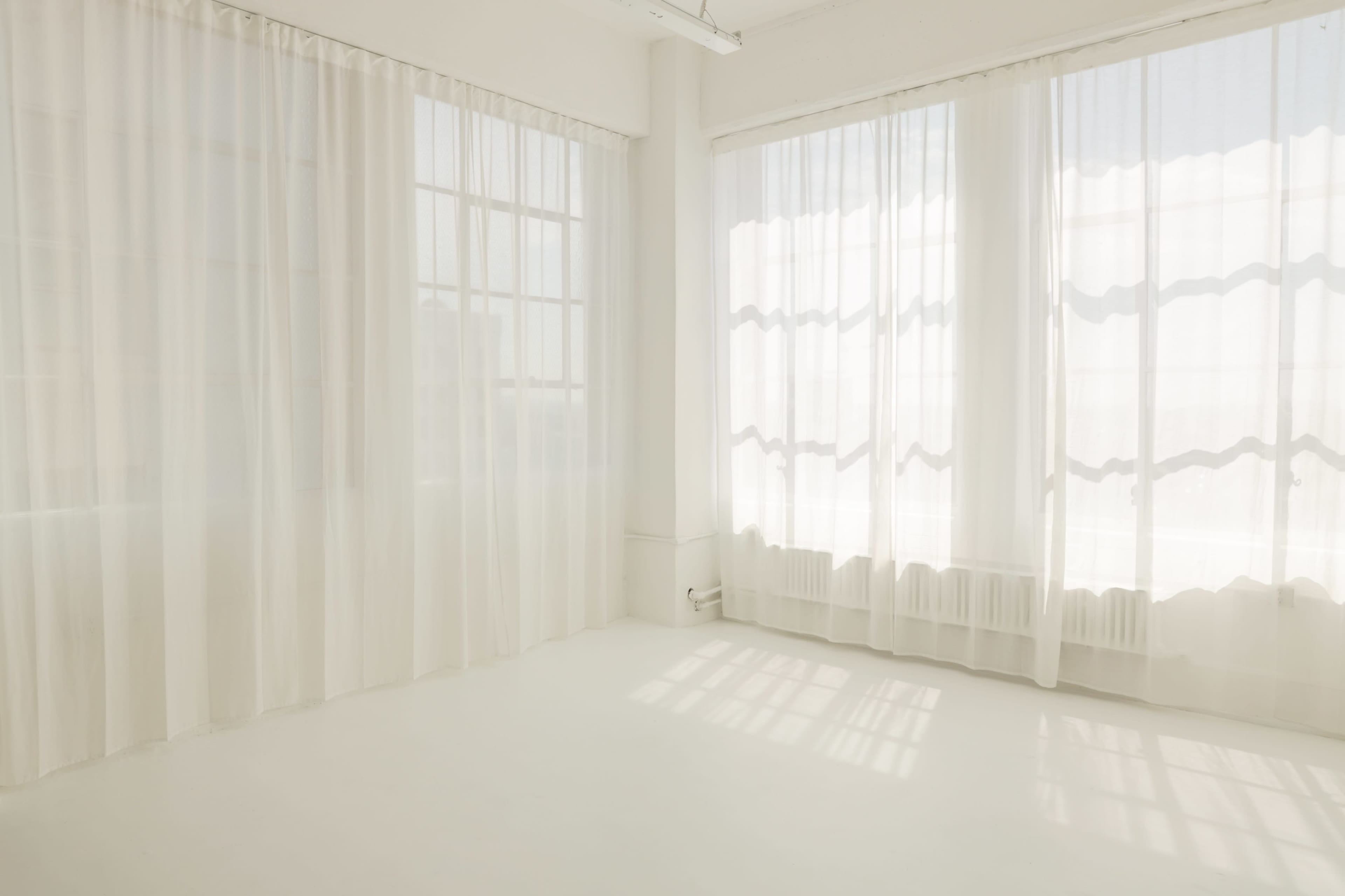 A bright, empty room with large windows covered by sheer white curtains, allowing soft light to filter in.