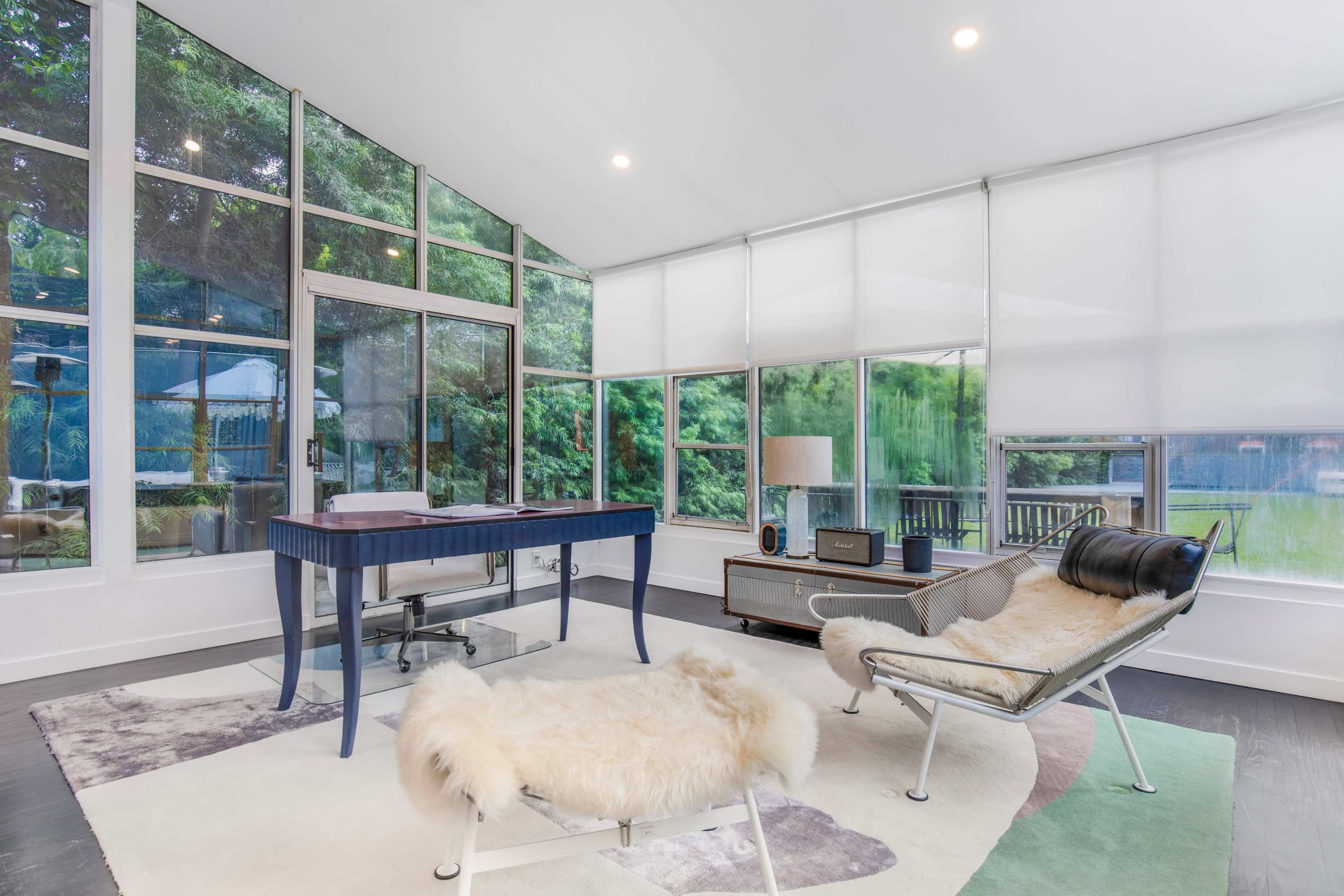 The image shows a modern workspace with a blue desk and a lounge chair, featuring large windows that provide a view of greenery outside.