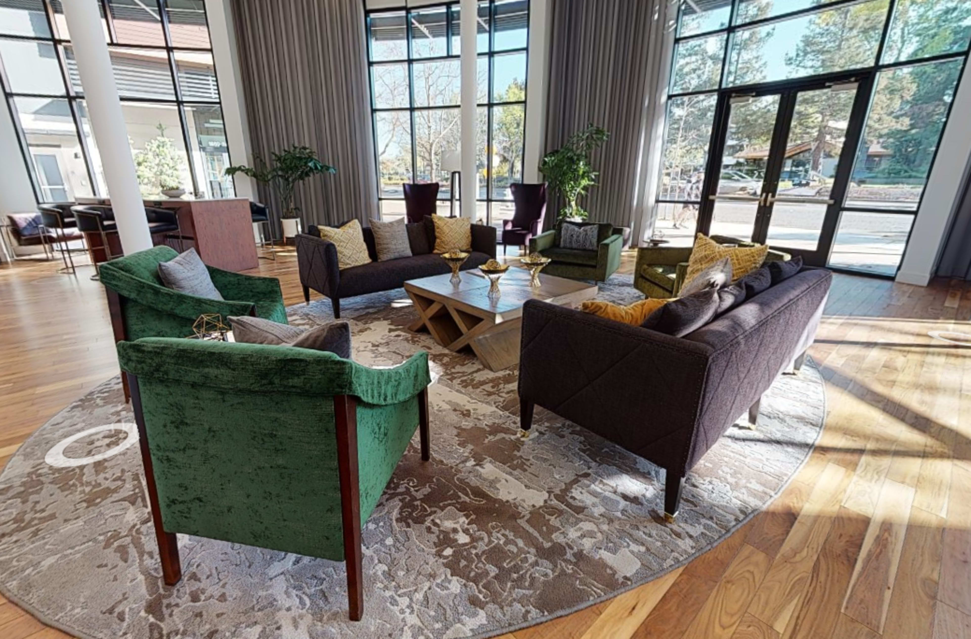A spacious lounge area featuring a circular rug, several upholstered chairs in green and dark brown, a coffee table, and large windows letting in natural light.
