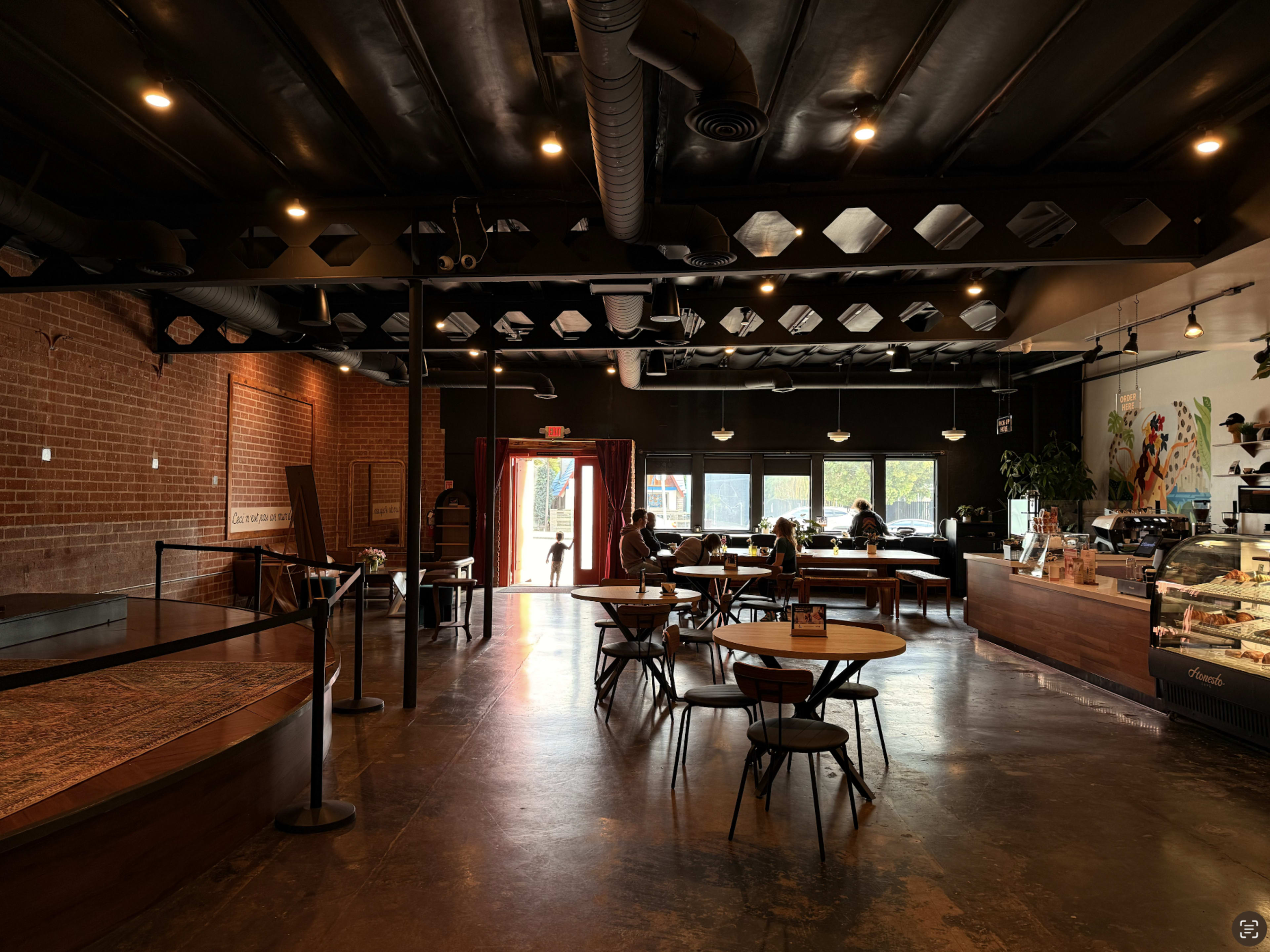 The image shows a modern coffee shop interior featuring wooden tables and chairs, with patrons seated and a counter displaying baked goods and beverages.