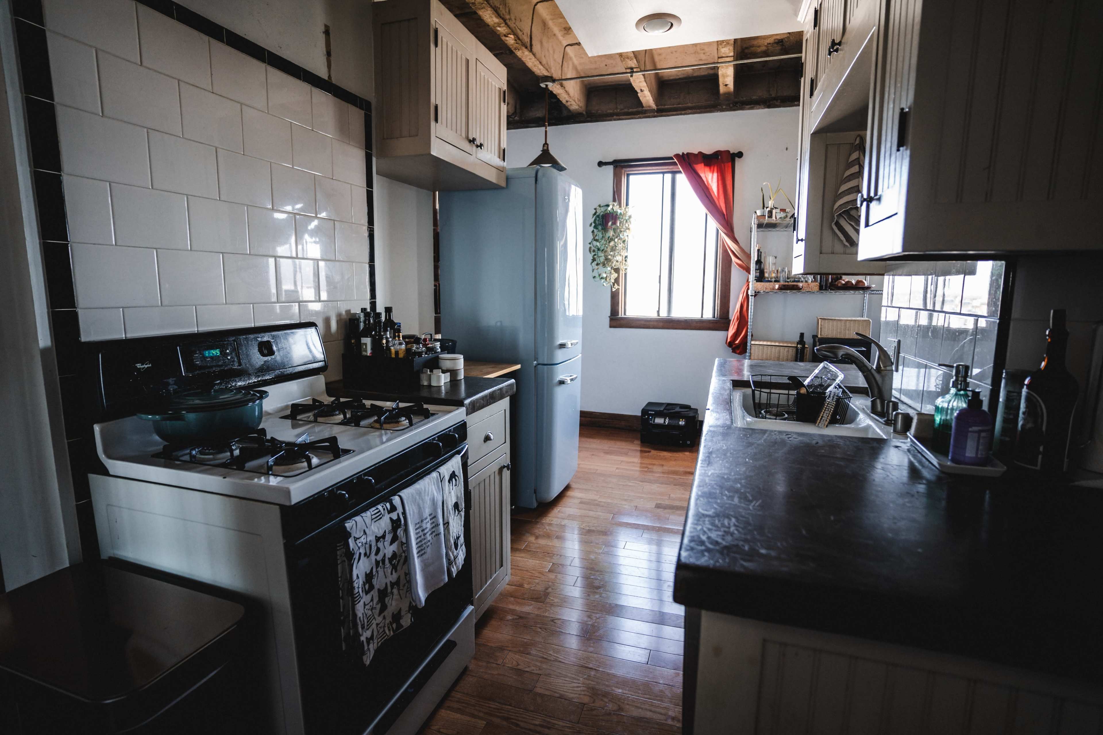 The image shows a kitchen featuring white cabinetry, a blue refrigerator, a gas stove, and wooden flooring, with a window dressed in red curtains.