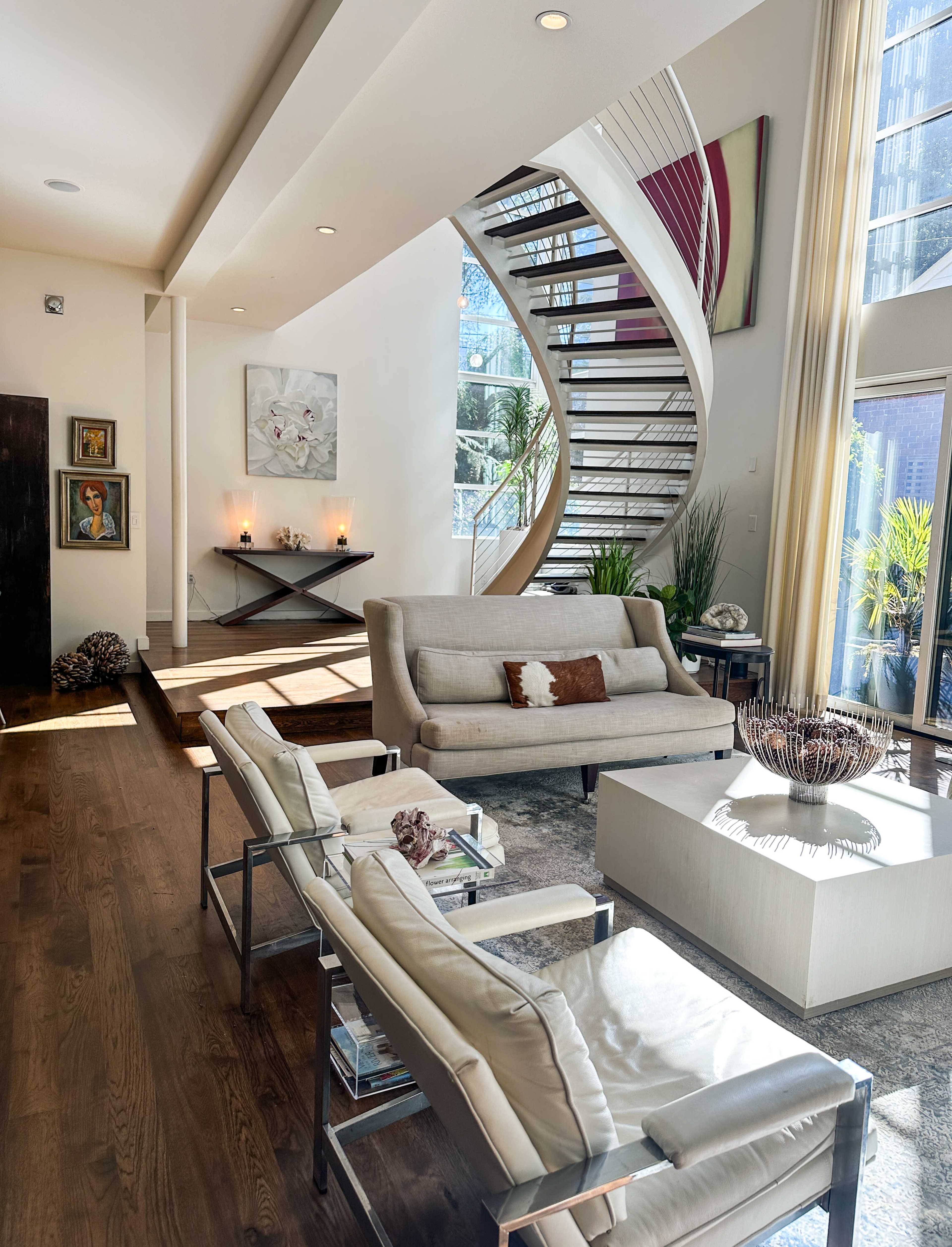 The image shows a modern living room with a spiral staircase, contemporary furniture, and large windows allowing natural light to enter.