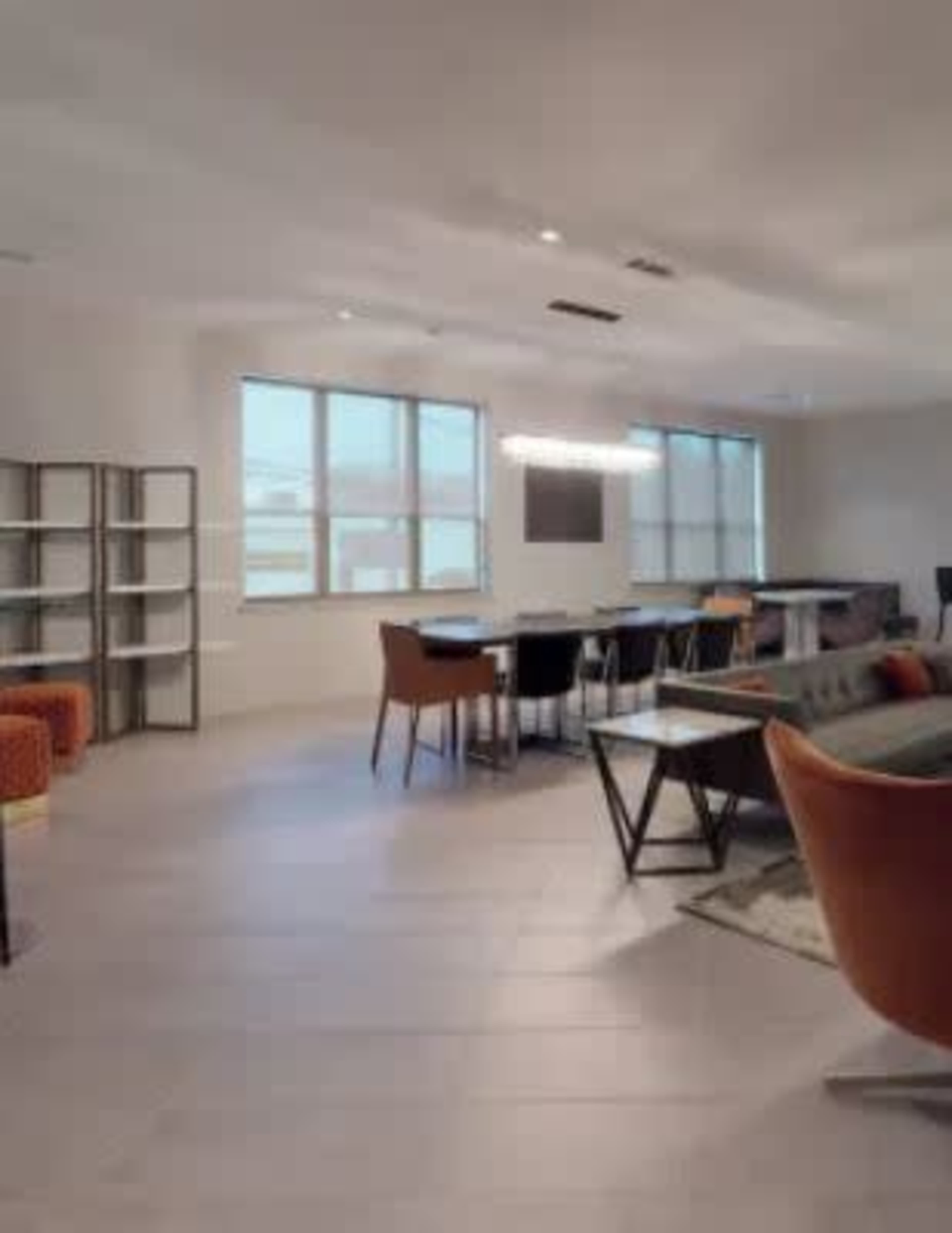 A modern lounge area with large windows, tables, chairs, and shelving units arranged in an open space.