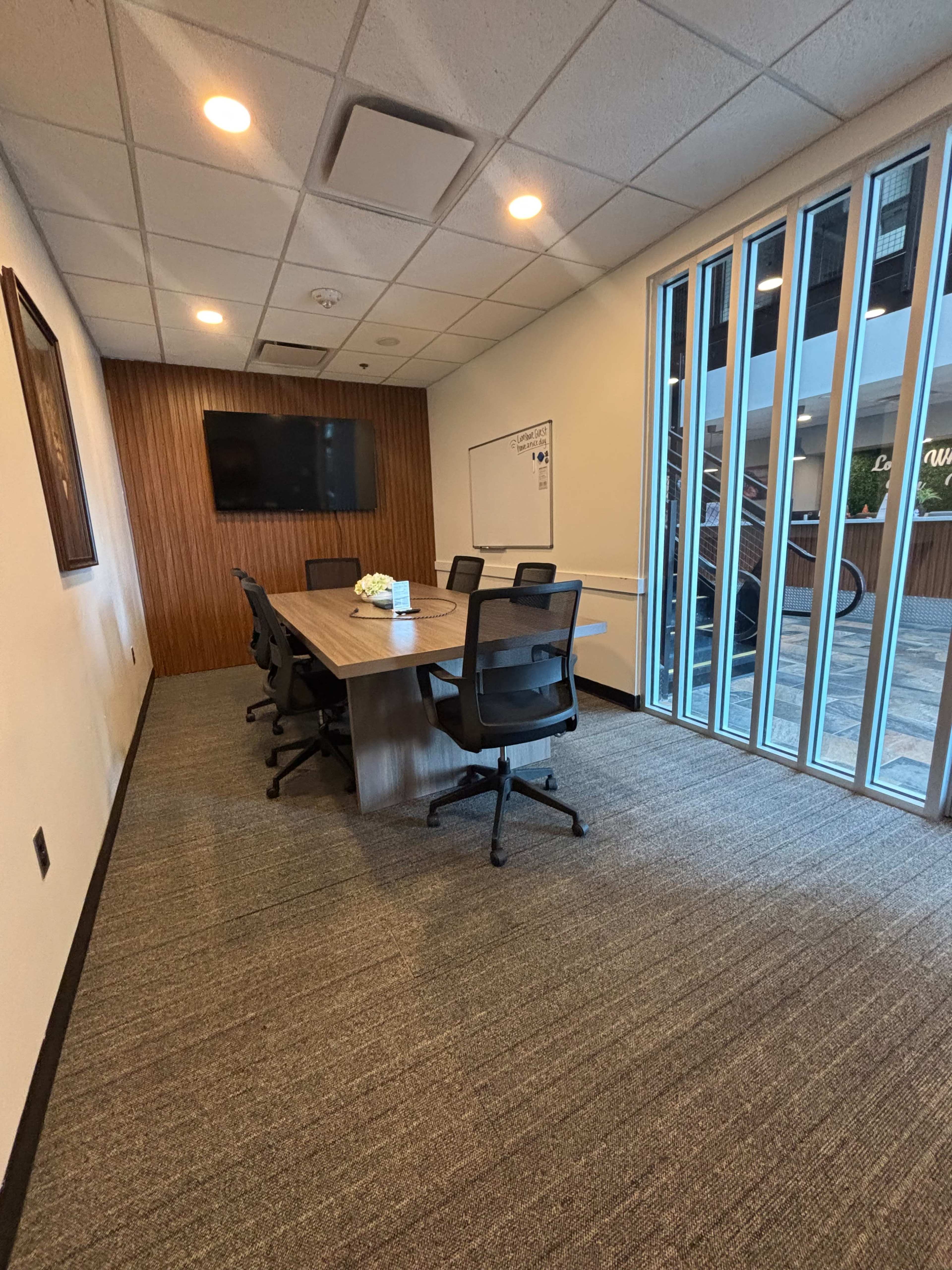 The image shows a conference room featuring a long table surrounded by six office chairs, with a whiteboard on one wall and large glass windows allowing natural light.