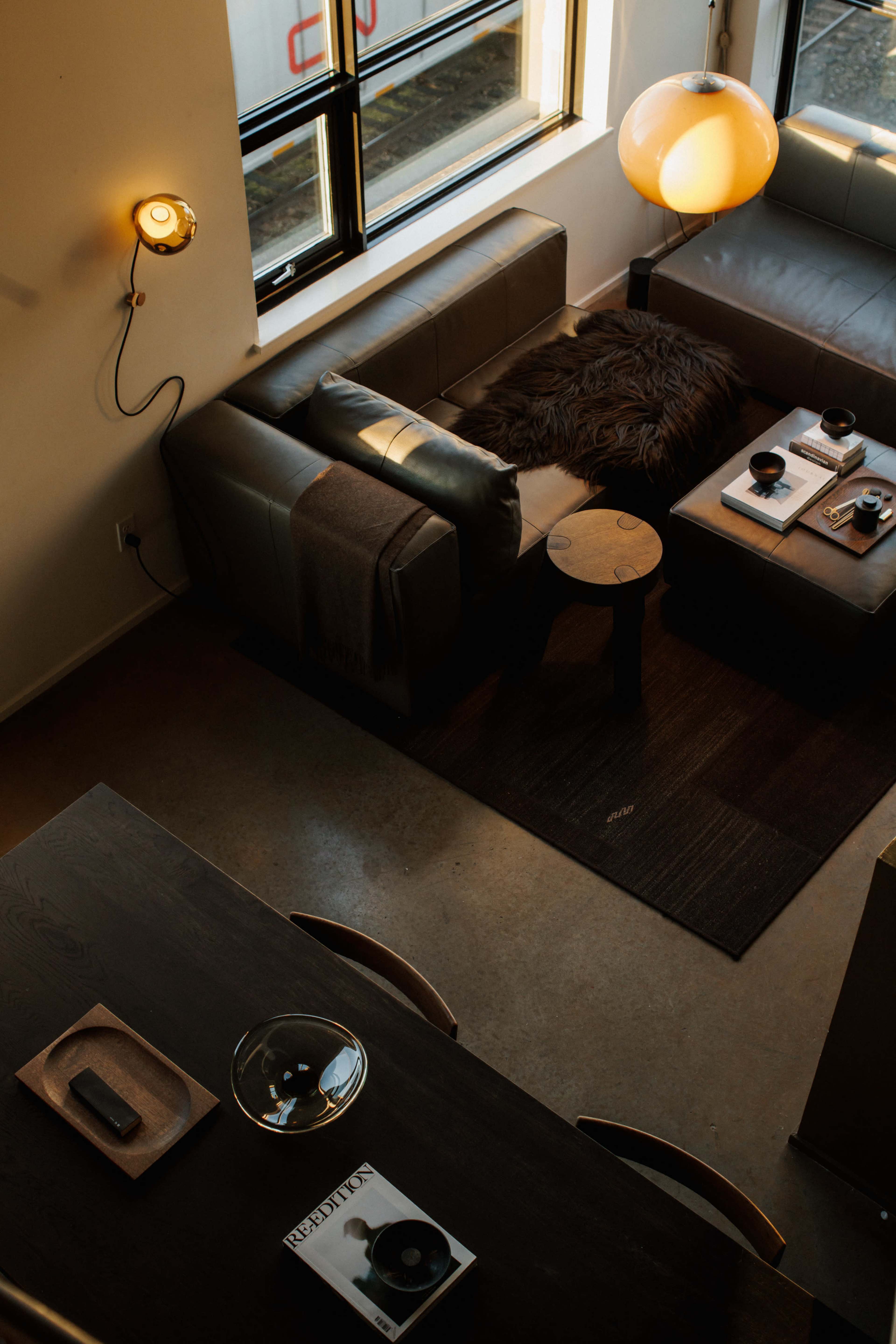 The image shows a cozy living area with leather sofas, a wooden coffee table, and a round lamp, illuminated by natural light coming through large windows.