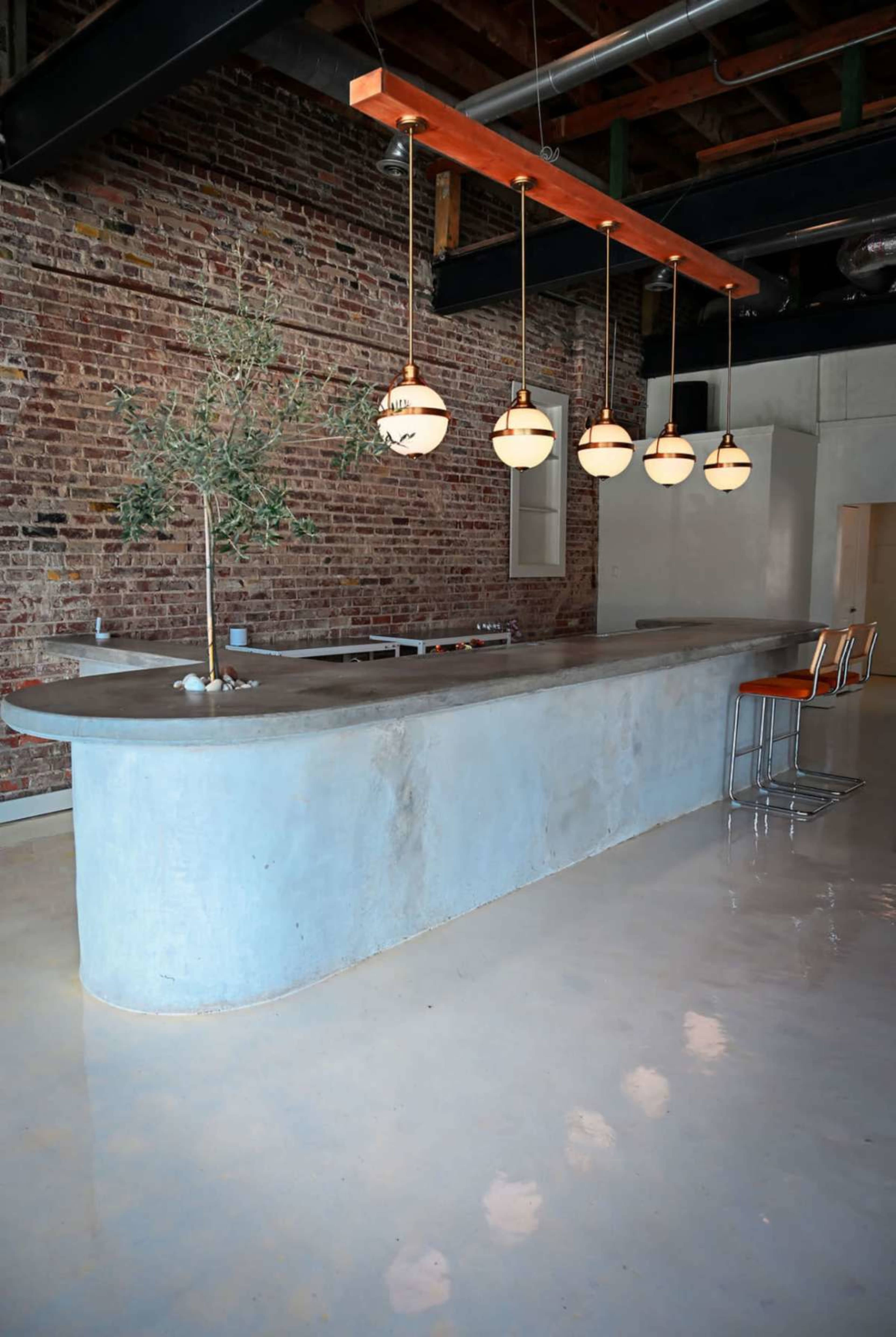 A curved concrete bar with pendant lights hangs above it, set against a backdrop of exposed brick walls and a polished floor.