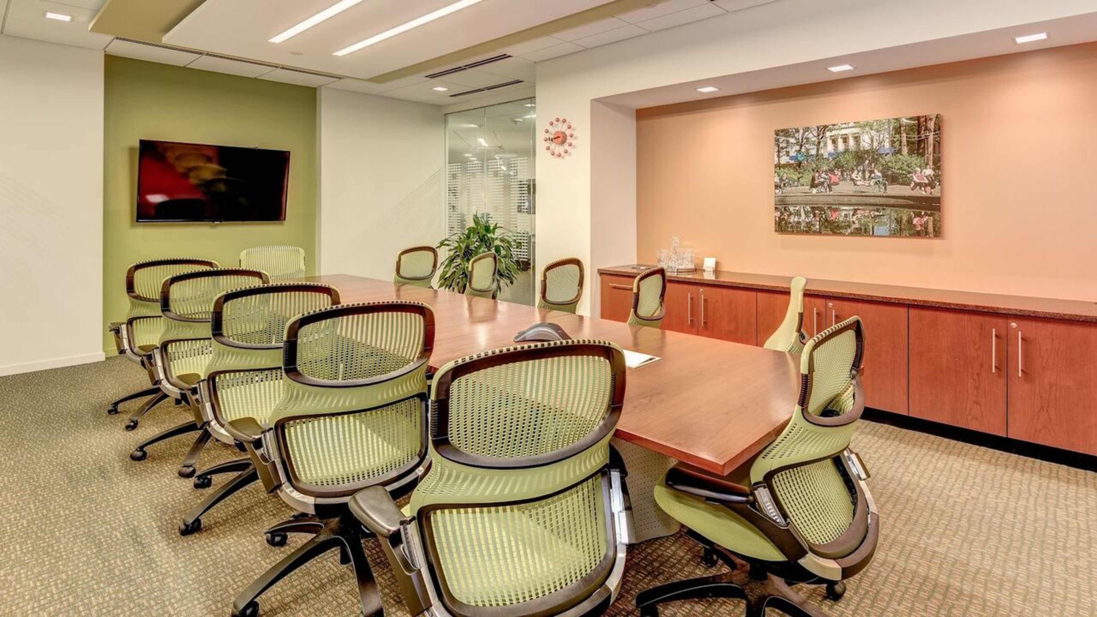 A modern conference room features a long wooden table surrounded by green ergonomic chairs, with a large screen and artwork on the walls.