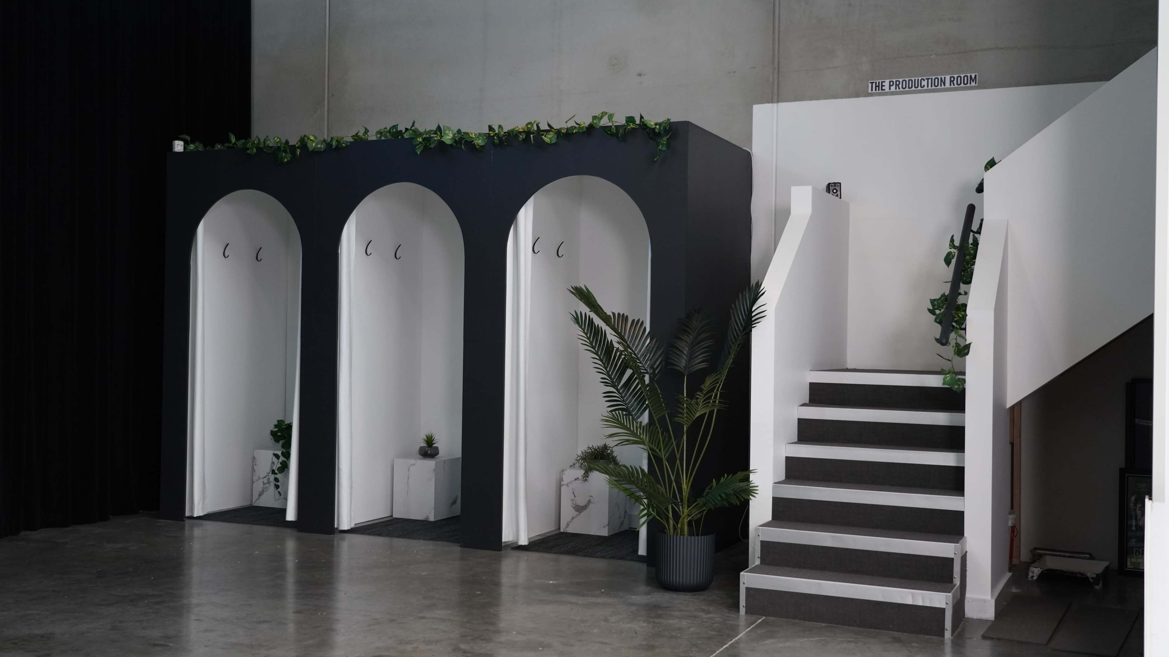 The image shows a modern interior featuring three arched alcoves with hooks, surrounded by greenery, alongside a staircase leading to another room labeled "THE PRODUCTION ROOM."