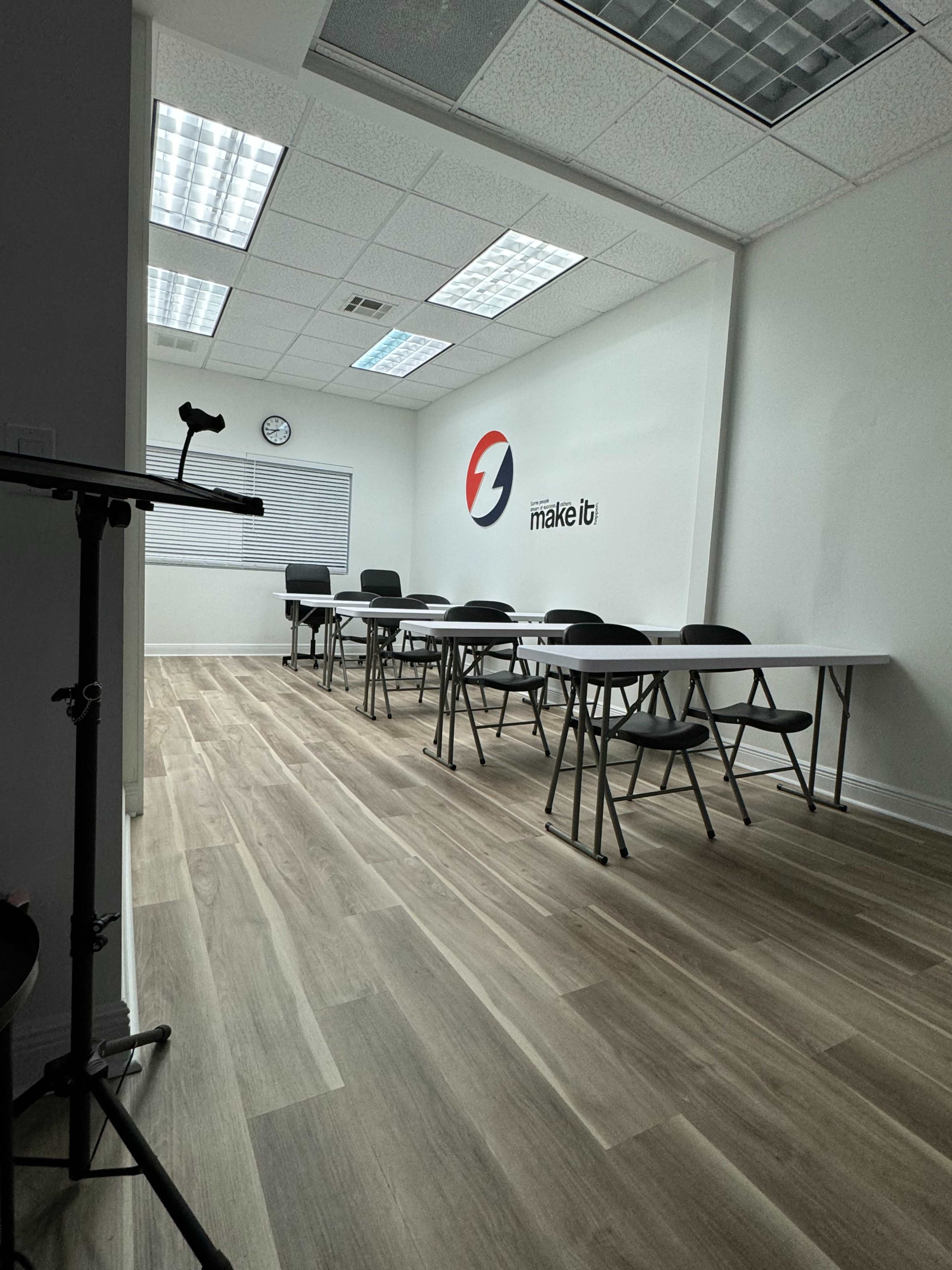 The image shows a spacious classroom with several rows of tables and chairs against a plain white wall, featuring a logo on one side and a clock on the opposite wall.