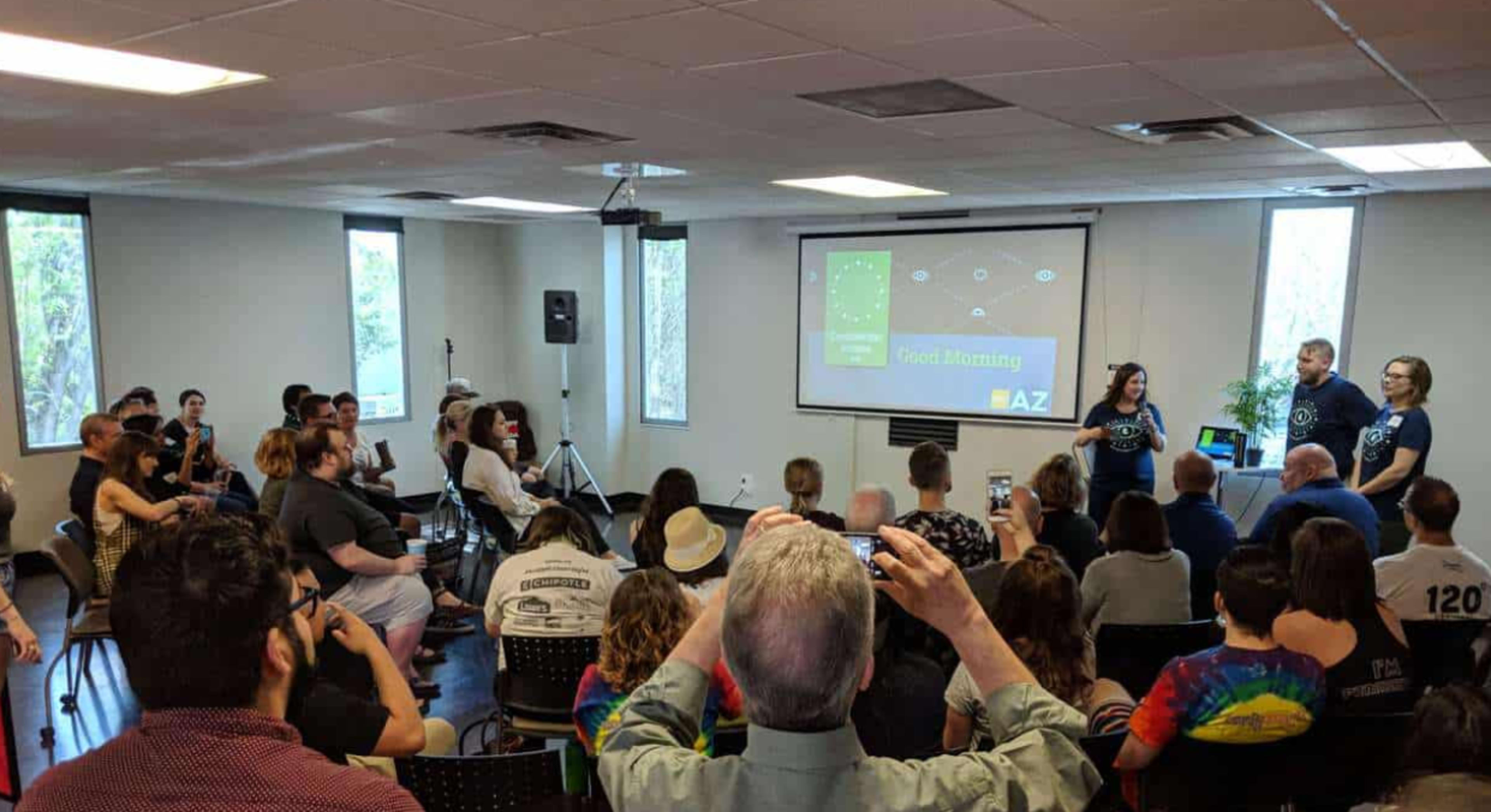 A group of people is seated in a conference room while two presenters speak at the front, with a large screen displaying the word "Good Morning" and a hashtag.