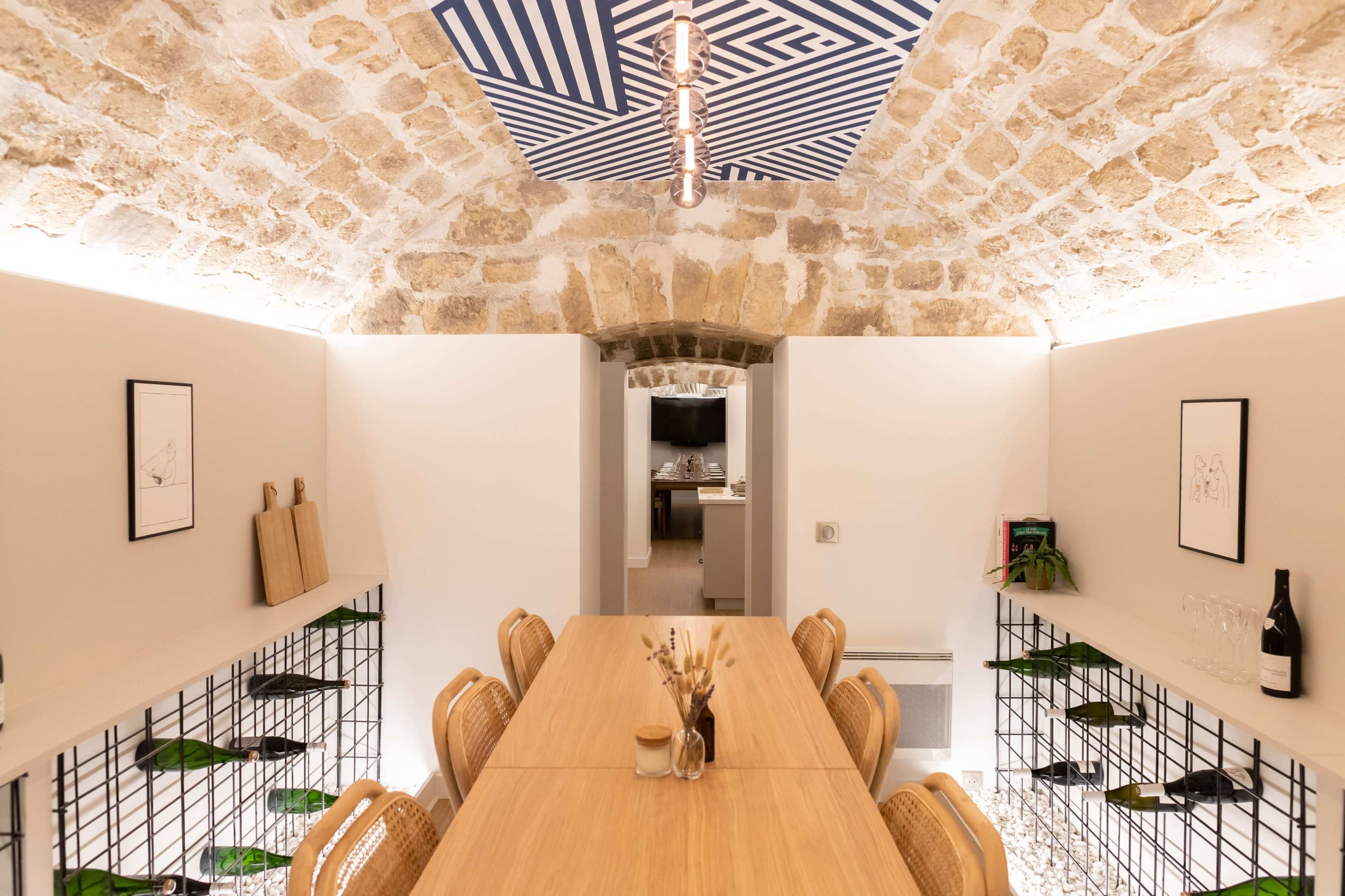 A dining area features a long wooden table surrounded by woven chairs, with a striped ceiling design and exposed stone walls.