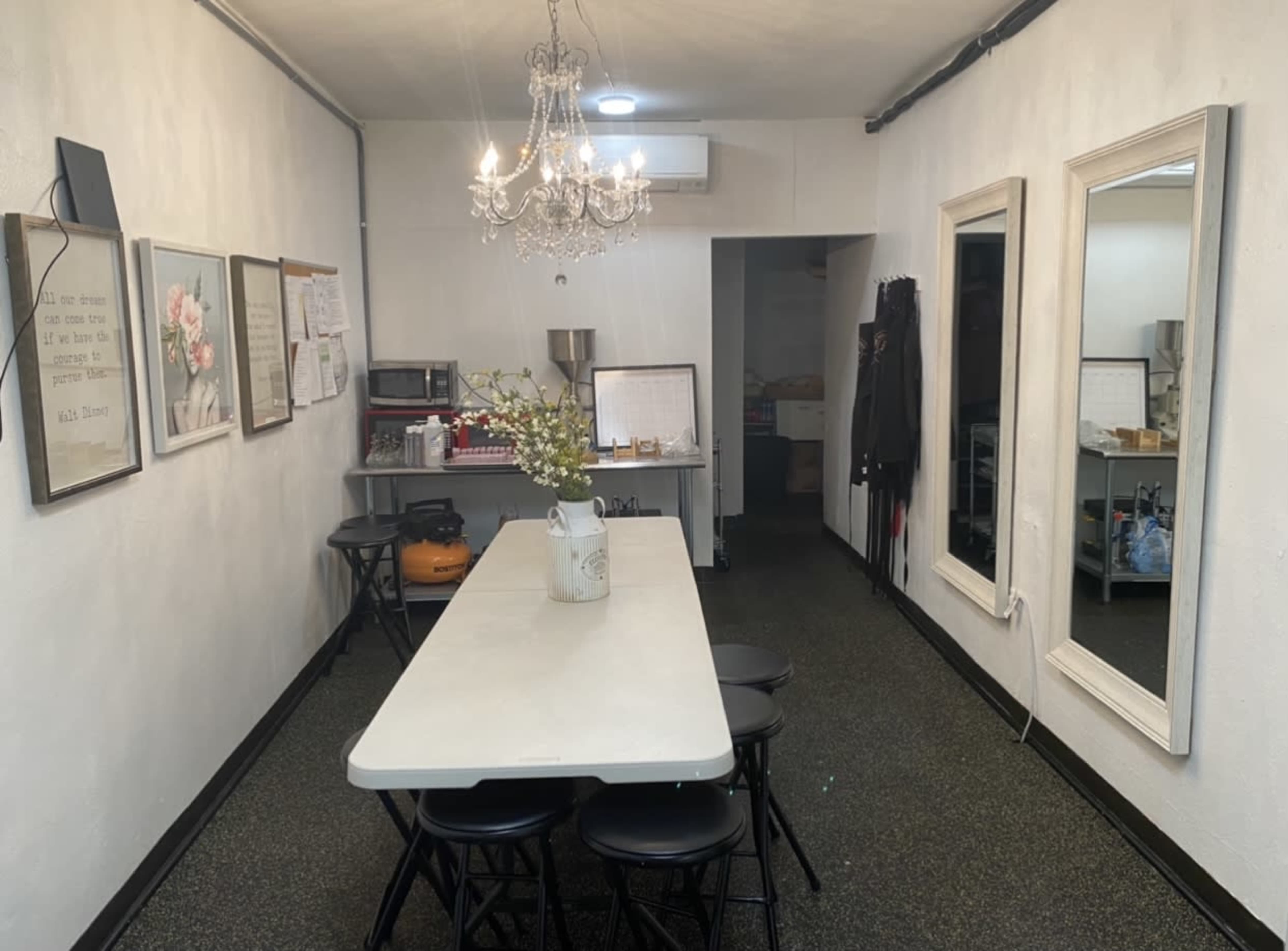 The image shows a simple dining area or meeting space with a long table, several black chairs, framed artwork on the walls, and a chandelier overhead.