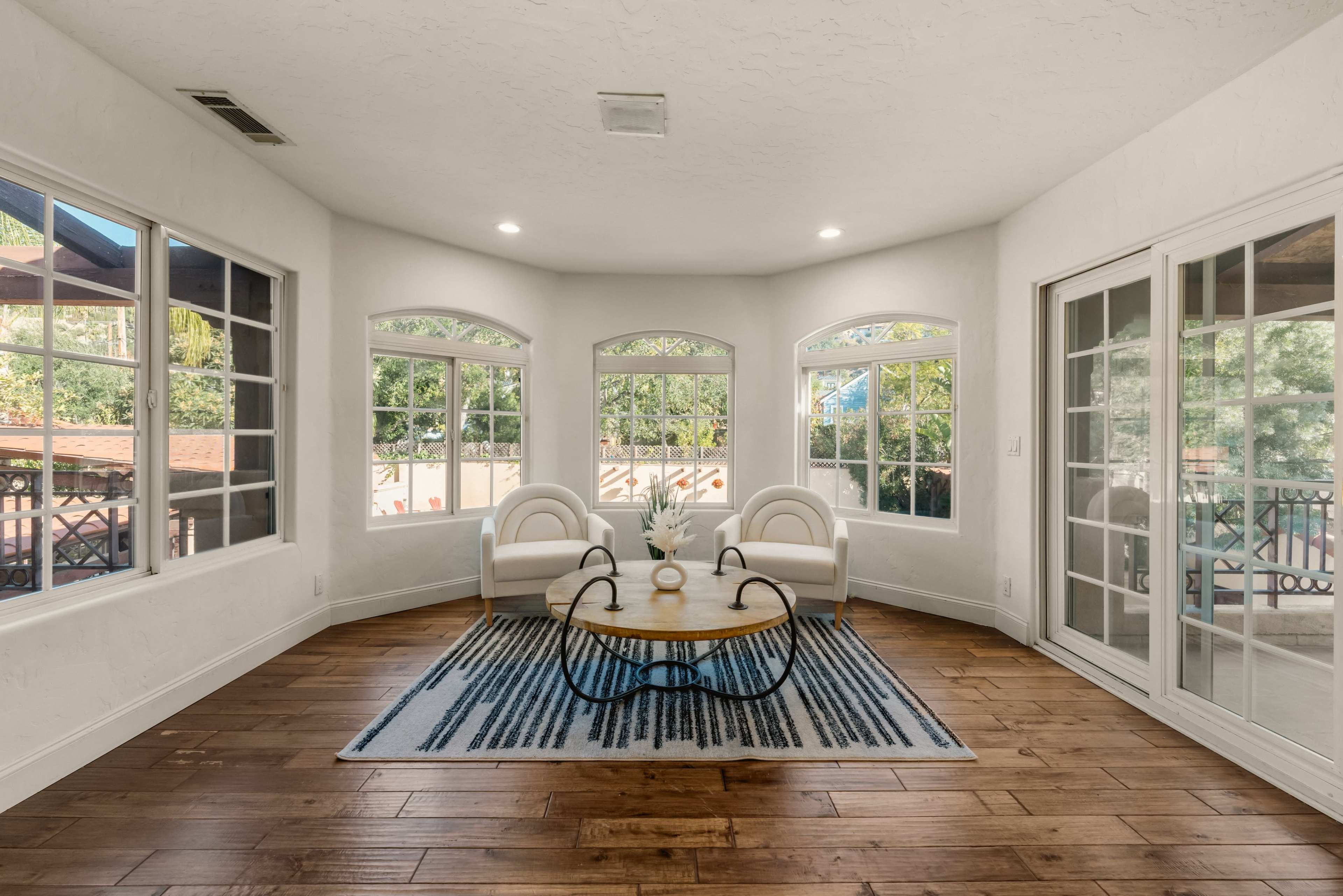 The image shows a bright room with large windows, two white chairs, and a wooden coffee table on a patterned rug, featuring hardwood floors.