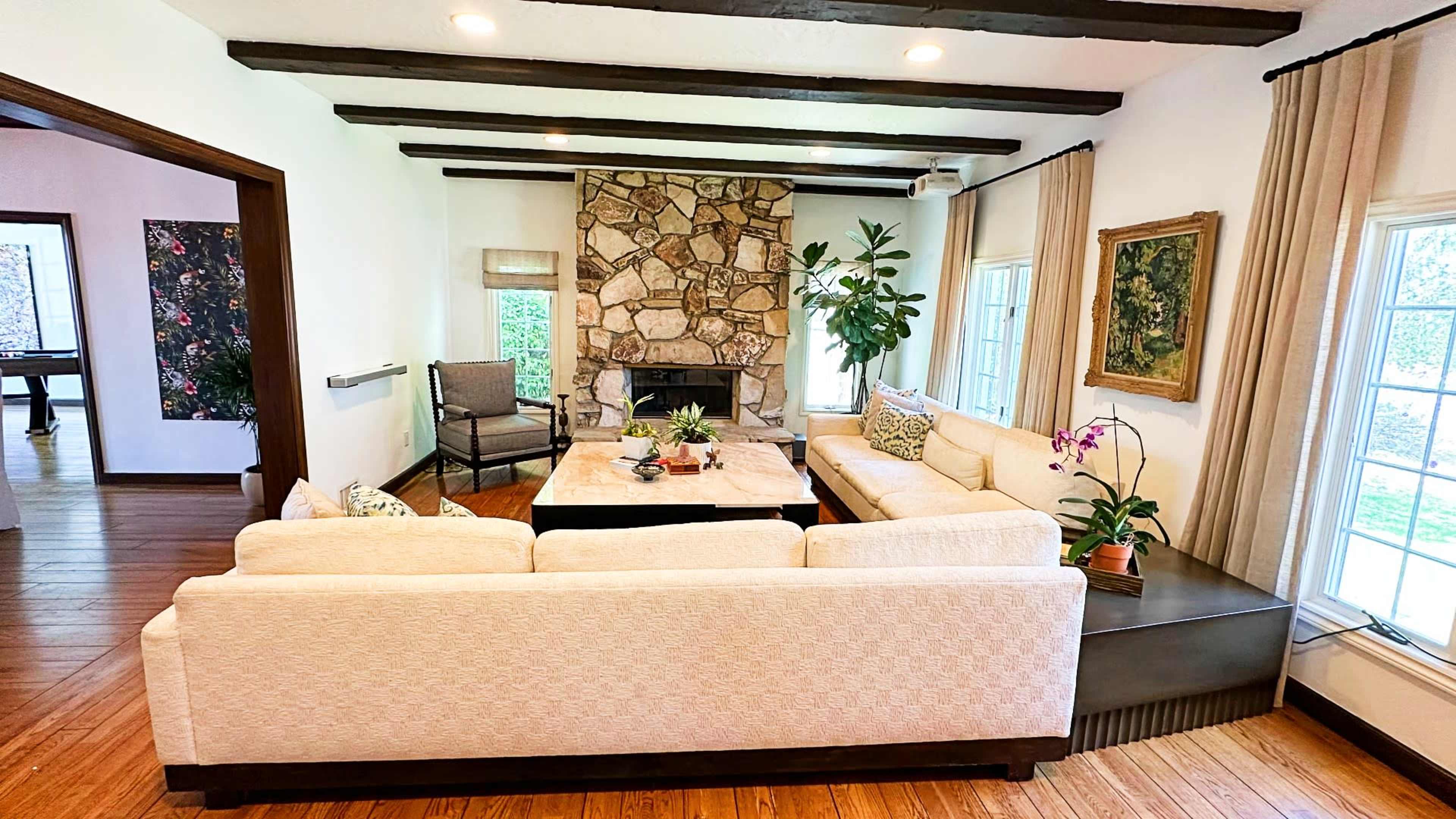 The living room features a large, light-colored sectional sofa facing a stone fireplace, with a coffee table at the center and windows providing natural light.