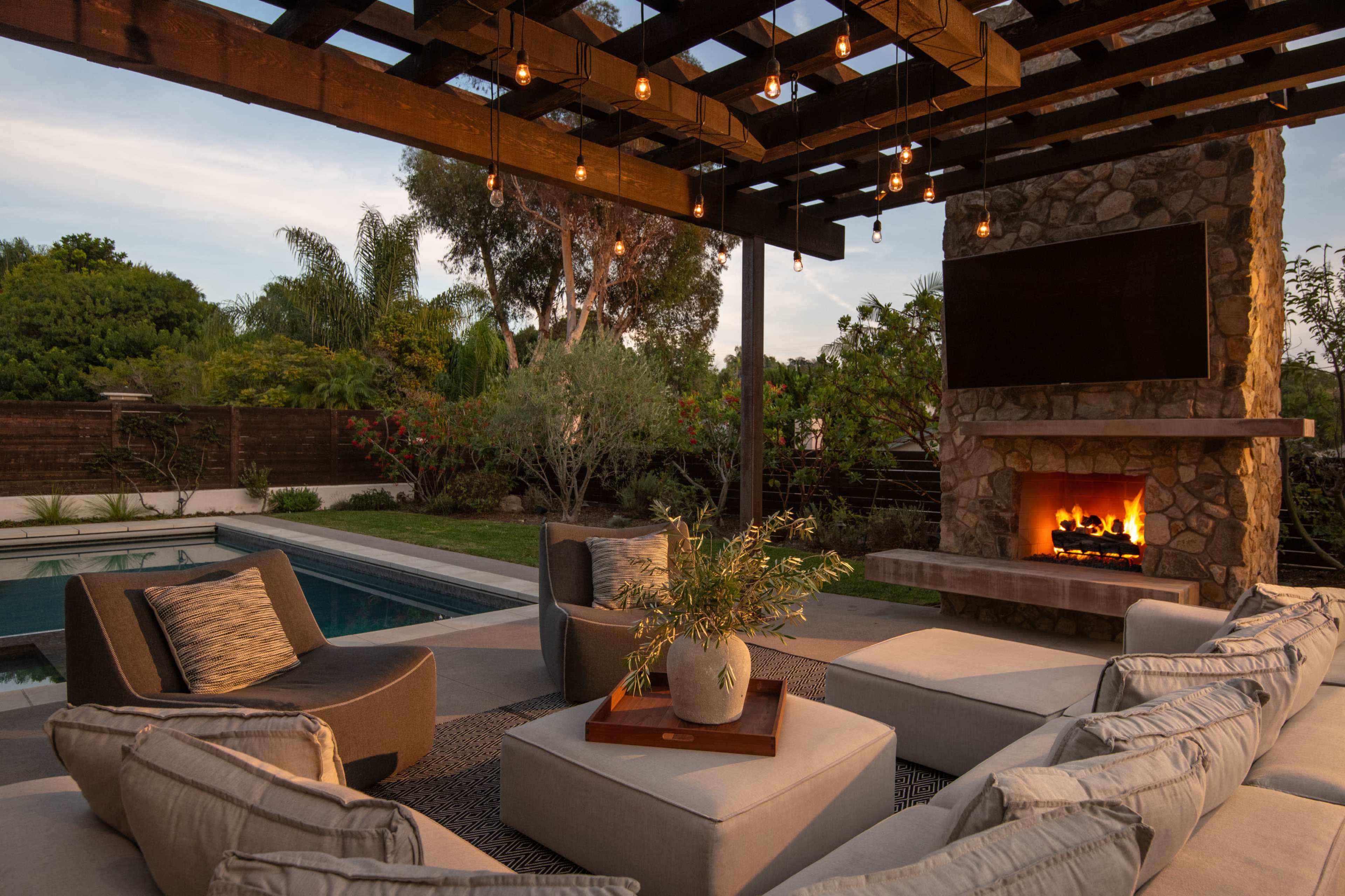 The image shows a cozy outdoor living area with a stone fireplace, modern seating, and a swimming pool surrounded by greenery.