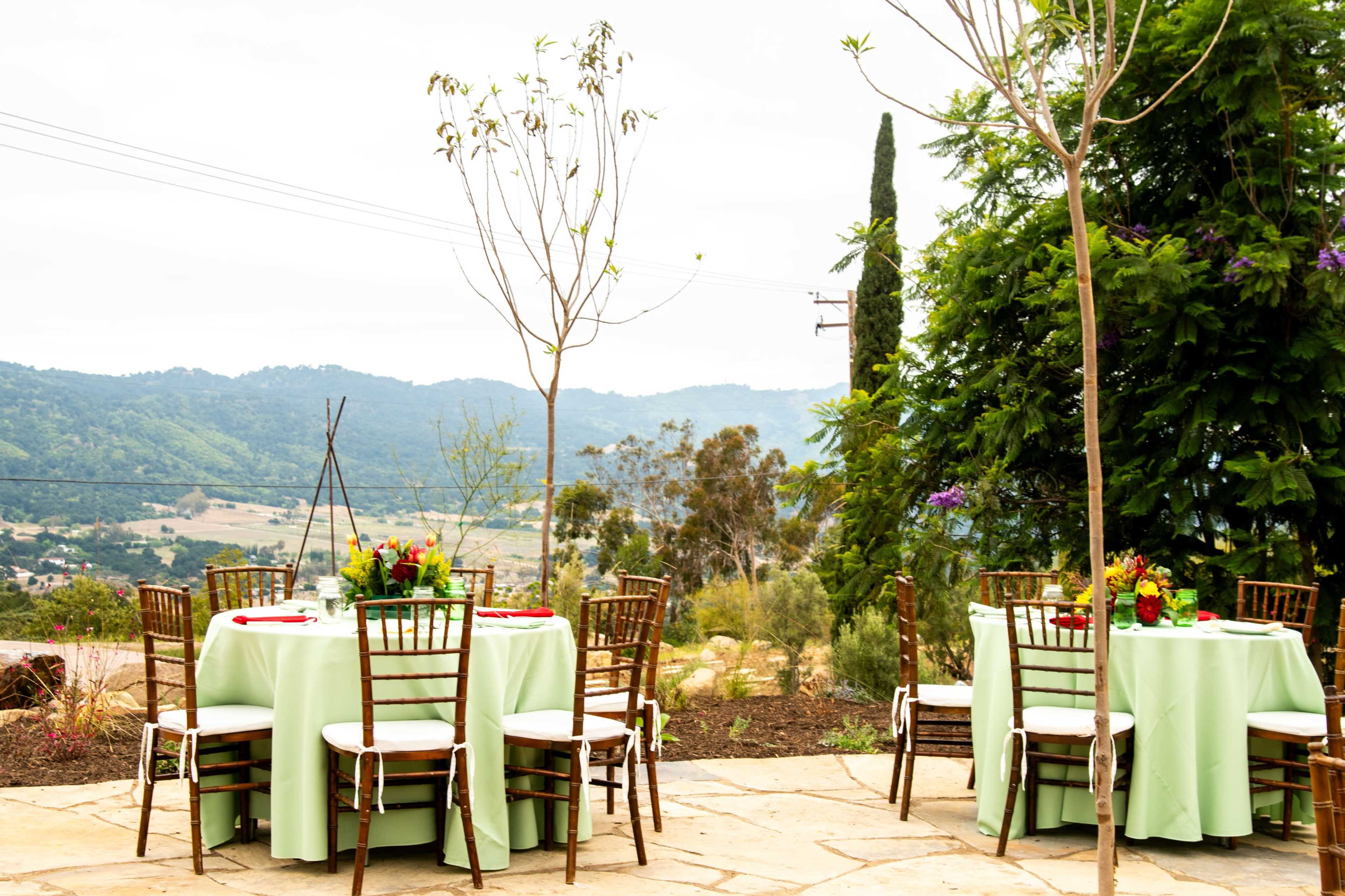 The image shows several elegantly set round tables with green tablecloths and floral centerpieces overlooking a scenic hillside with trees in the background.