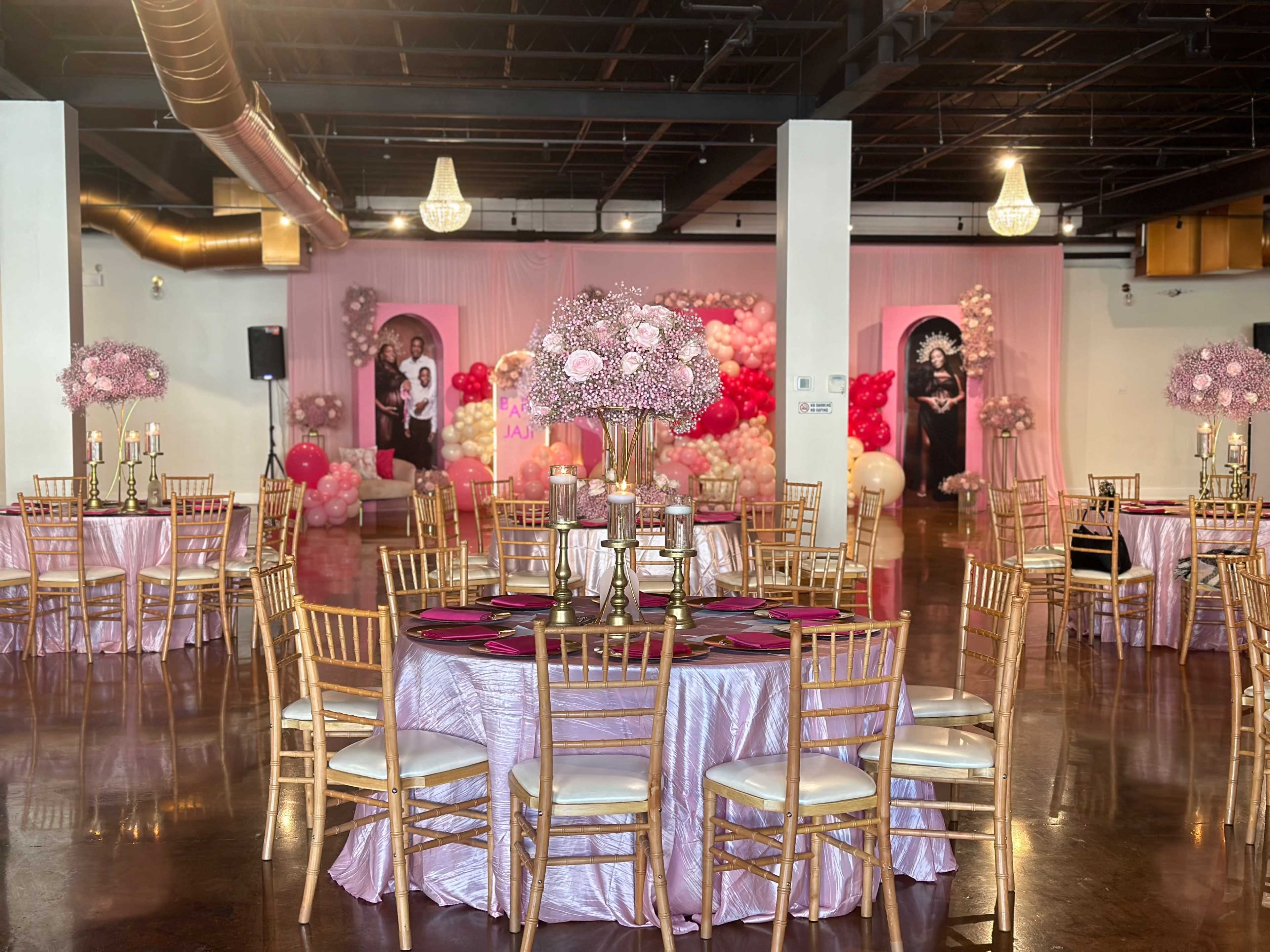 The image shows a decorated event space with round tables covered in pink tablecloths, gold chairs, and floral centerpieces, featuring a backdrop of balloons and portraits.