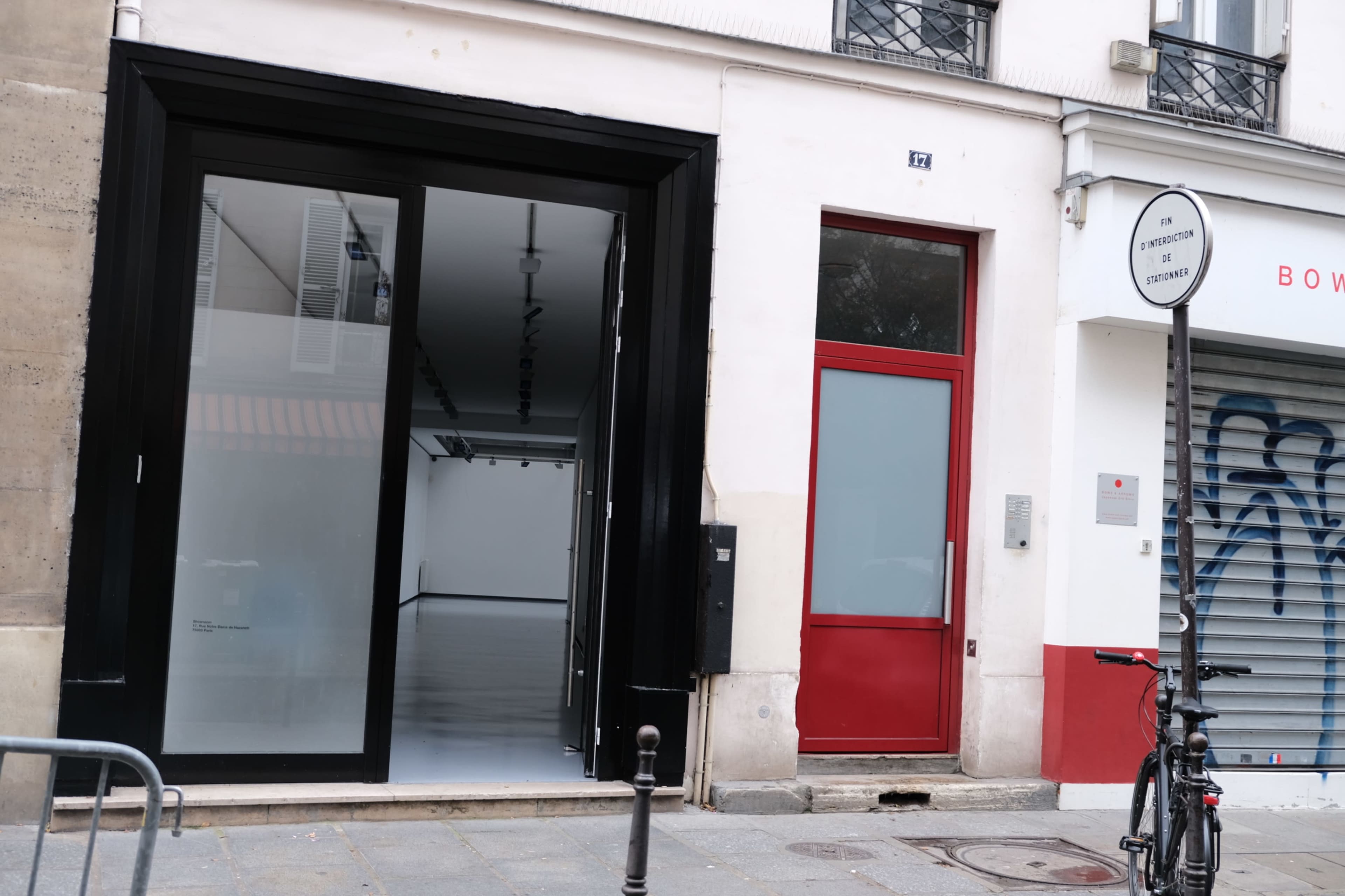 The image shows a minimalist storefront with a large black-framed glass entrance next to a red door, set on a city street with a parked bicycle nearby.